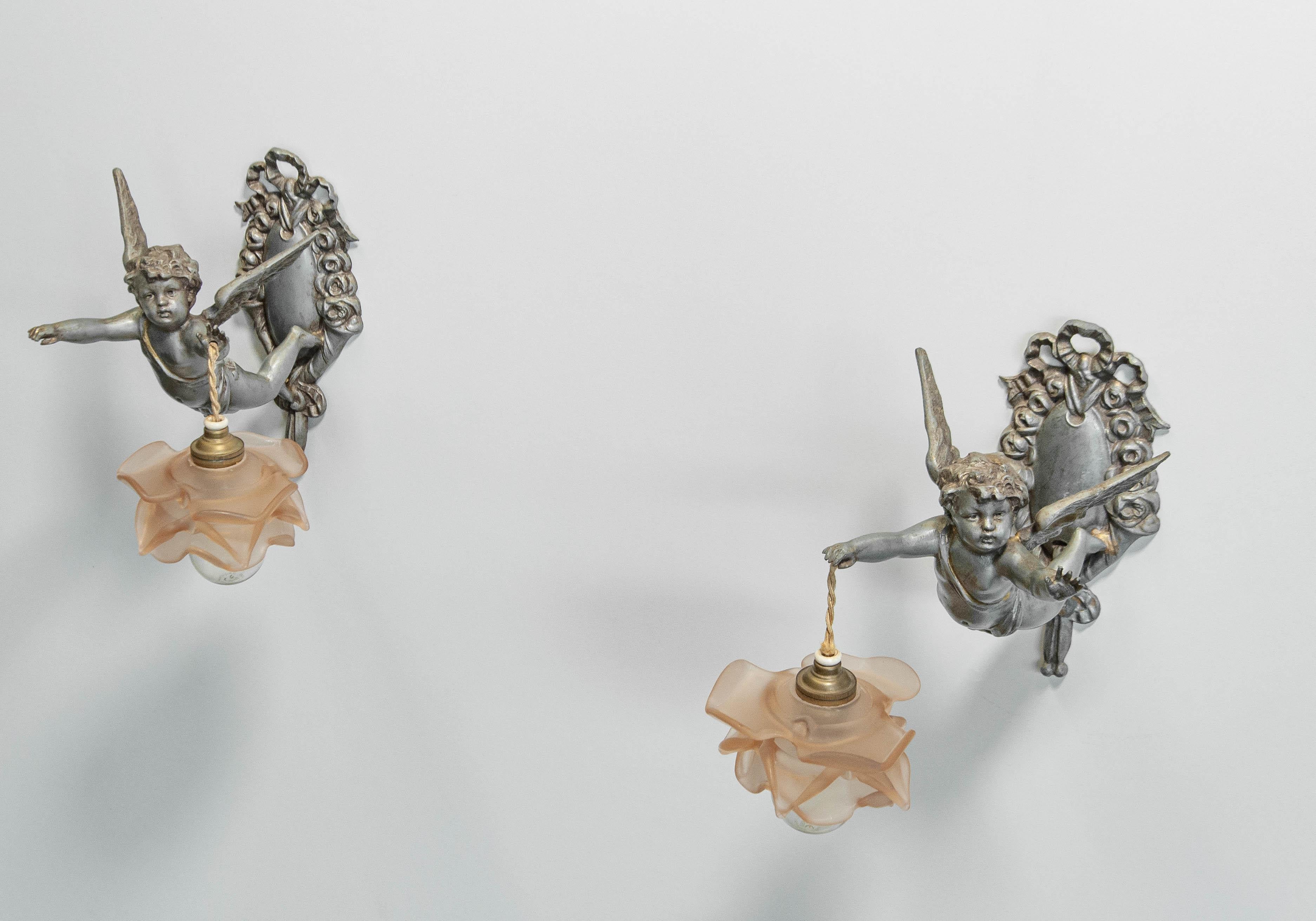 Rare matching pair of French Rococo-style wall sconces, crafted in pewter, dating from the 1920s. Each sconce features a finely detailed cherub holding a frosted art glass shade with soft pink ruffled edges. Retaining their original French fittings