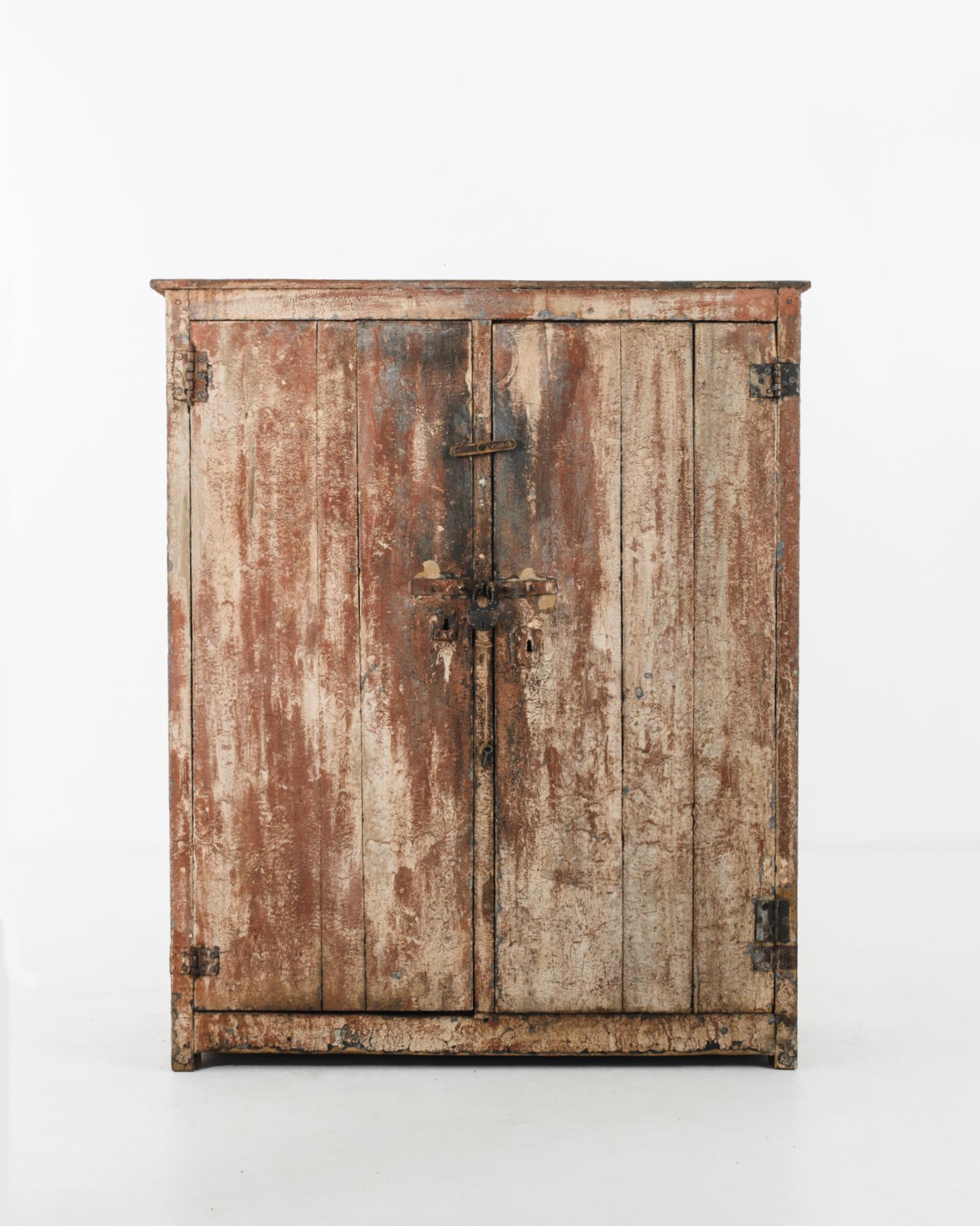 A wooden cabinet from 1920s Belgium, characterized by a distinctive patina. Two hinged doors set within a square case close with a cast iron lock; a rustic detail which complements the rawness of the patinated finish. Rust, ash and cream tones