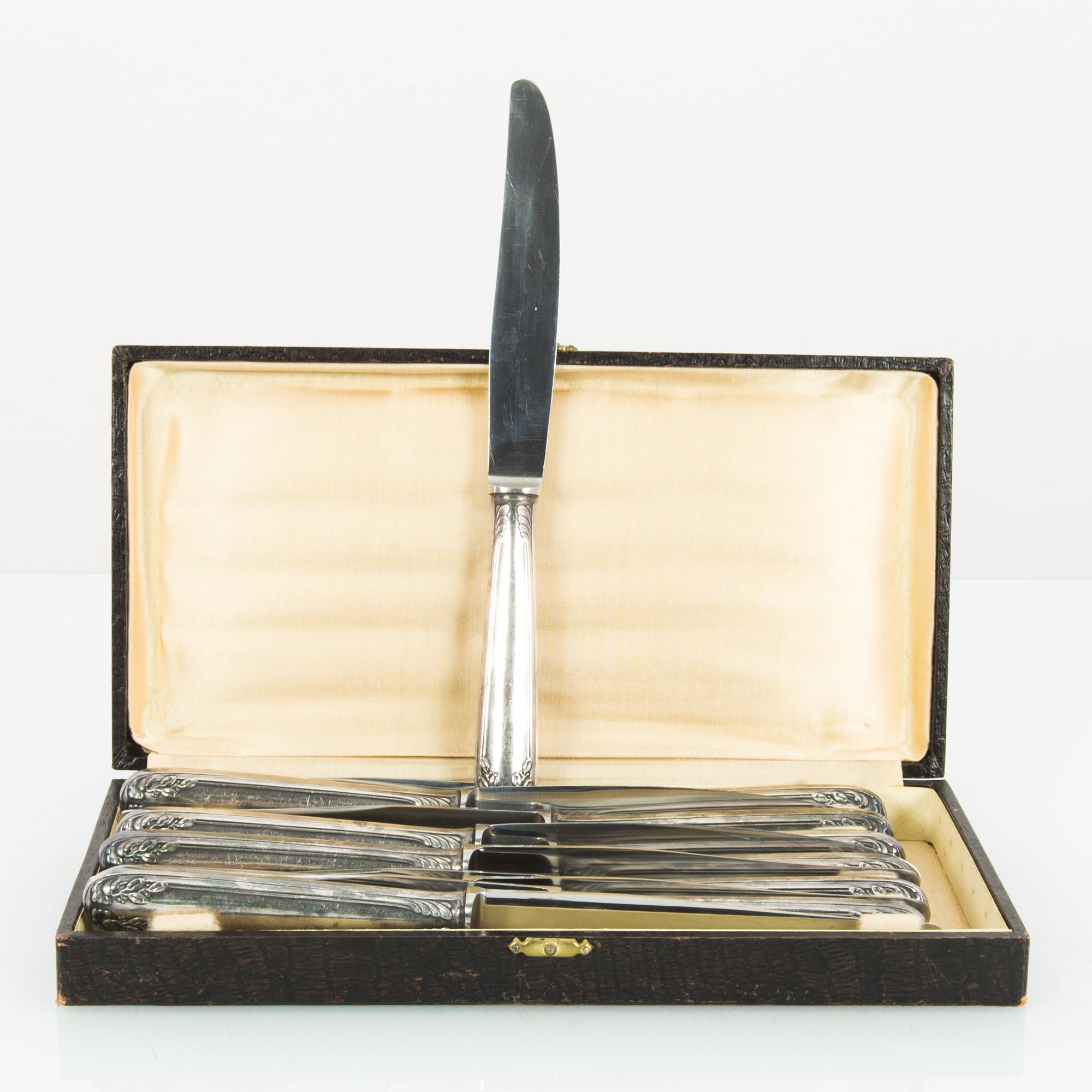 A set of silver-plated knives from Europe, circa 1920. Silver knives sit within a rectangular box, which closes with a gold clasp. The box is finished in dark brown snakeskin; the interior is lined with rose-gold silk. The handles of the knives are