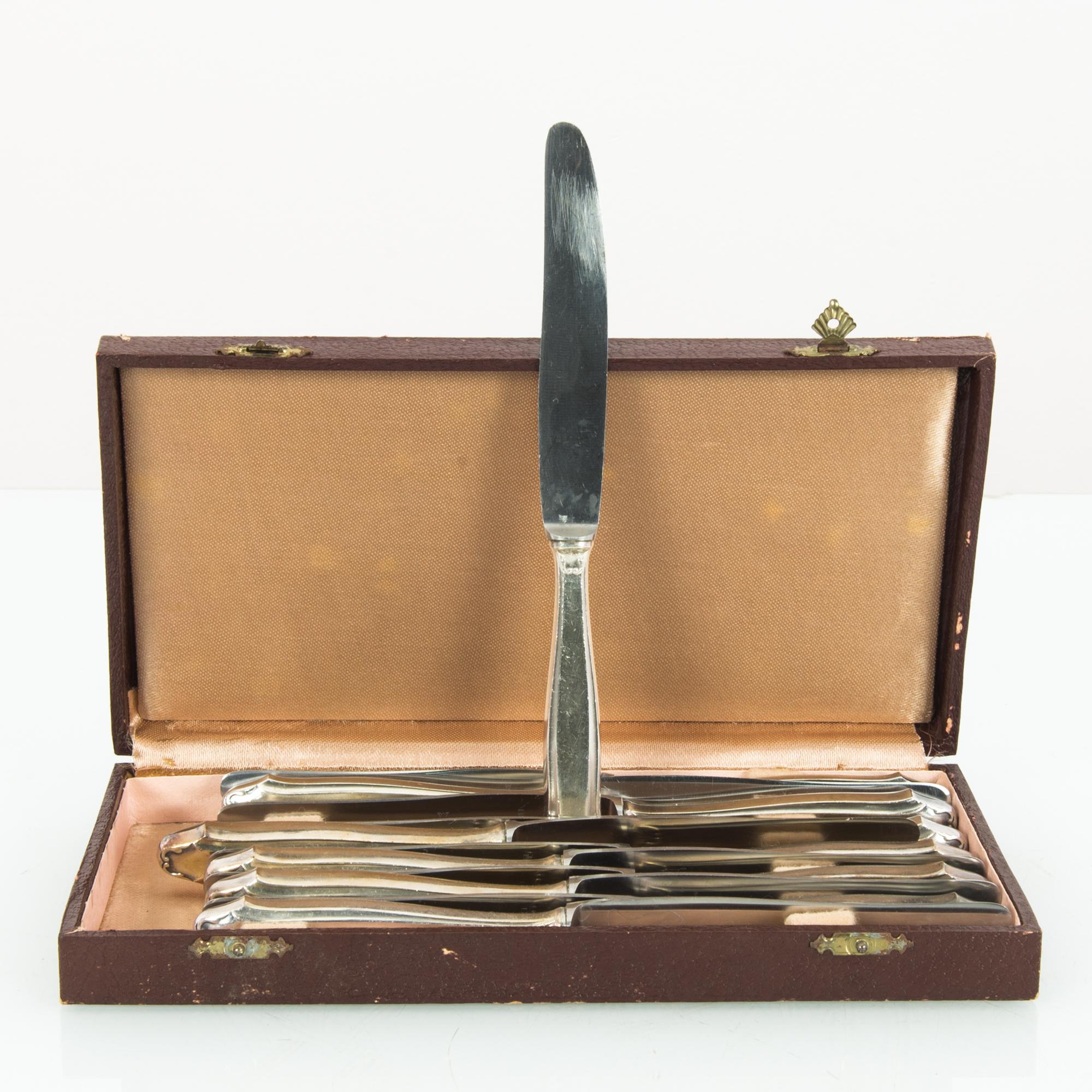 A set of silver-plated knives from Europe, circa 1920. Silver knives are poised within a rectangular box, which closes with a pair of gilded shell-shaped clasps. The exterior is finished in oxblood leather; the inside is lined with soft peach silk.