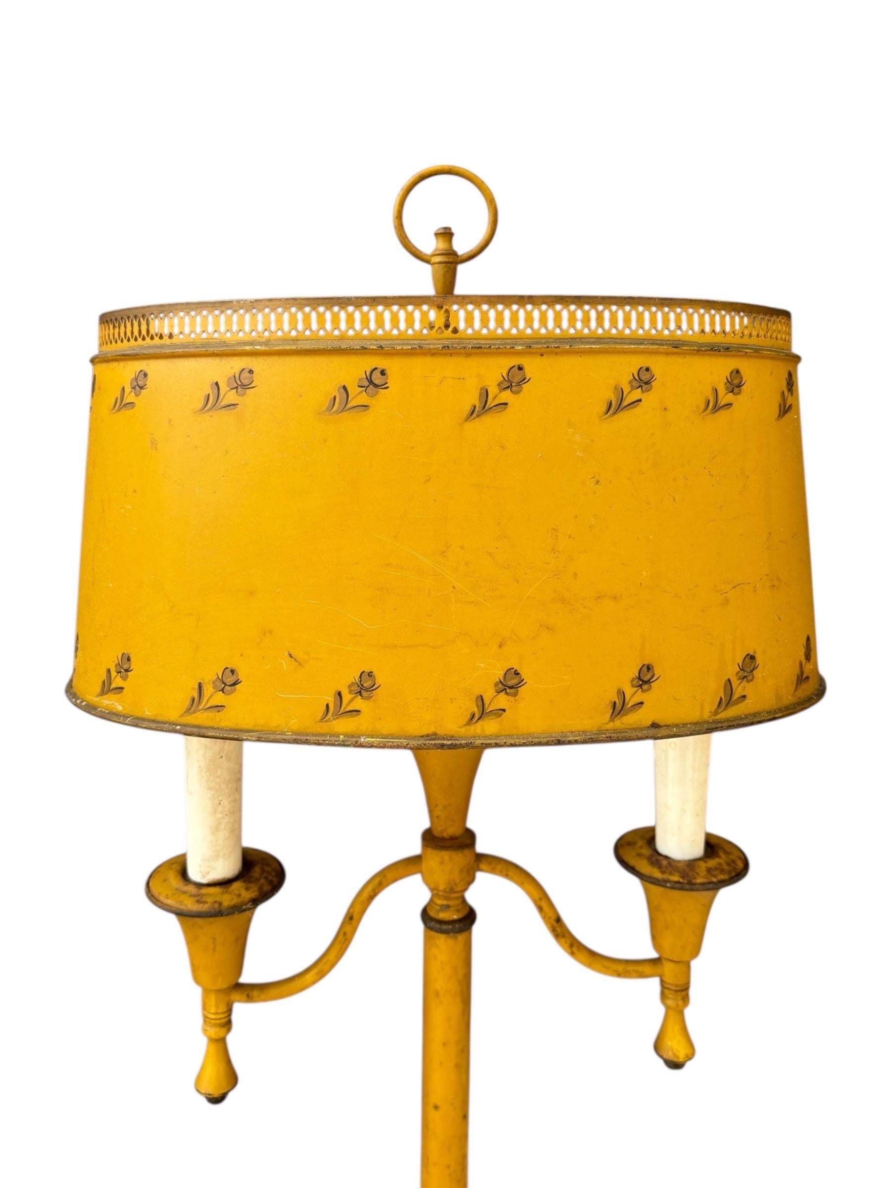 Una llamativa lámpara de mesa bouillotte de tole amarillo de hacia 1920. Una columna central sostiene dos ménsulas de brazos con sencillos capiteles cónicos. Arriba está la pantalla ovalada, decorada en la parte superior con una galería calada.