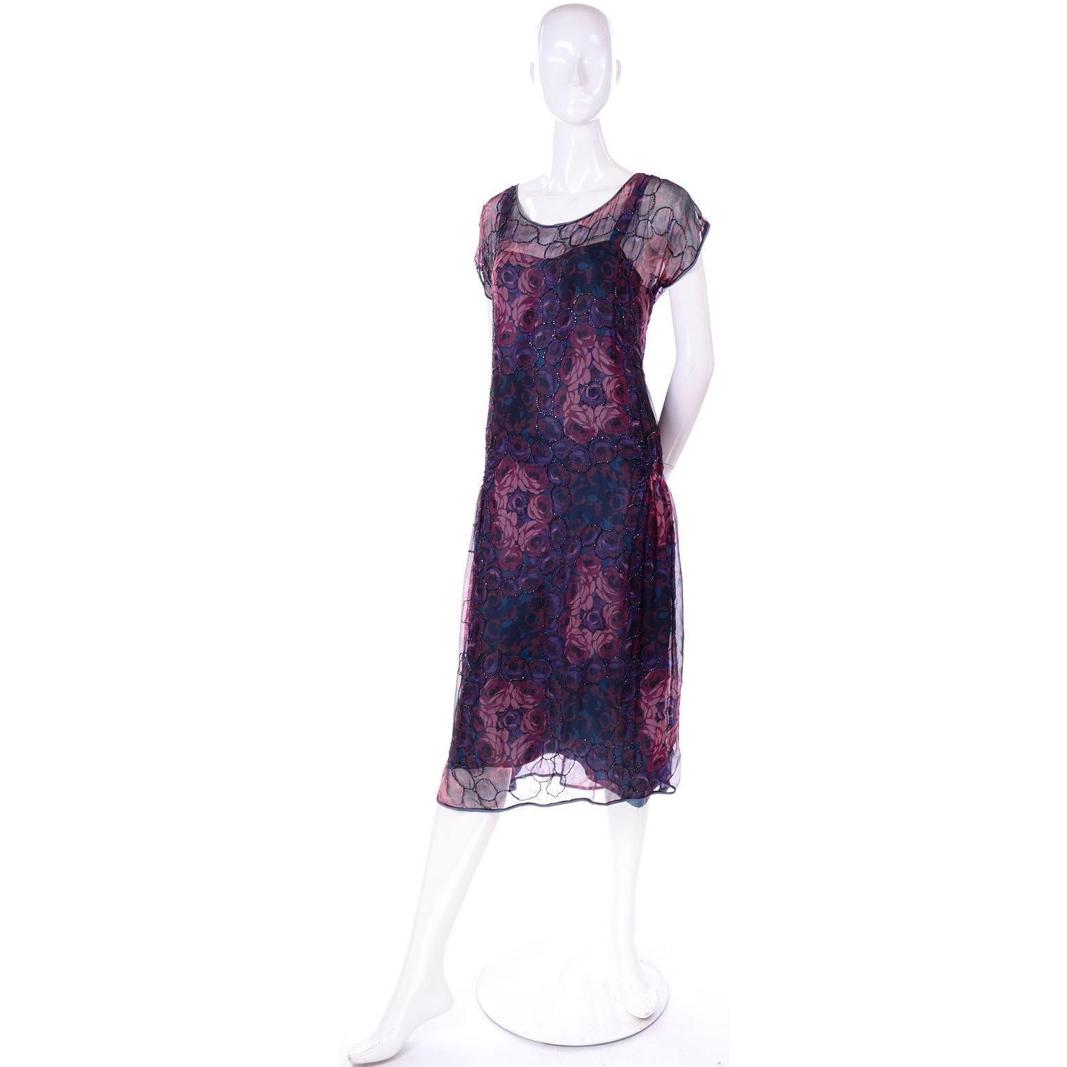 This is a lovely sheer floral 1920's dress with black faceted beading around swirls of pink and purple roses. This pretty 20's dress has pale blue trim and an attached blue silk slip under-dress that shows through the slit in the side of the outer