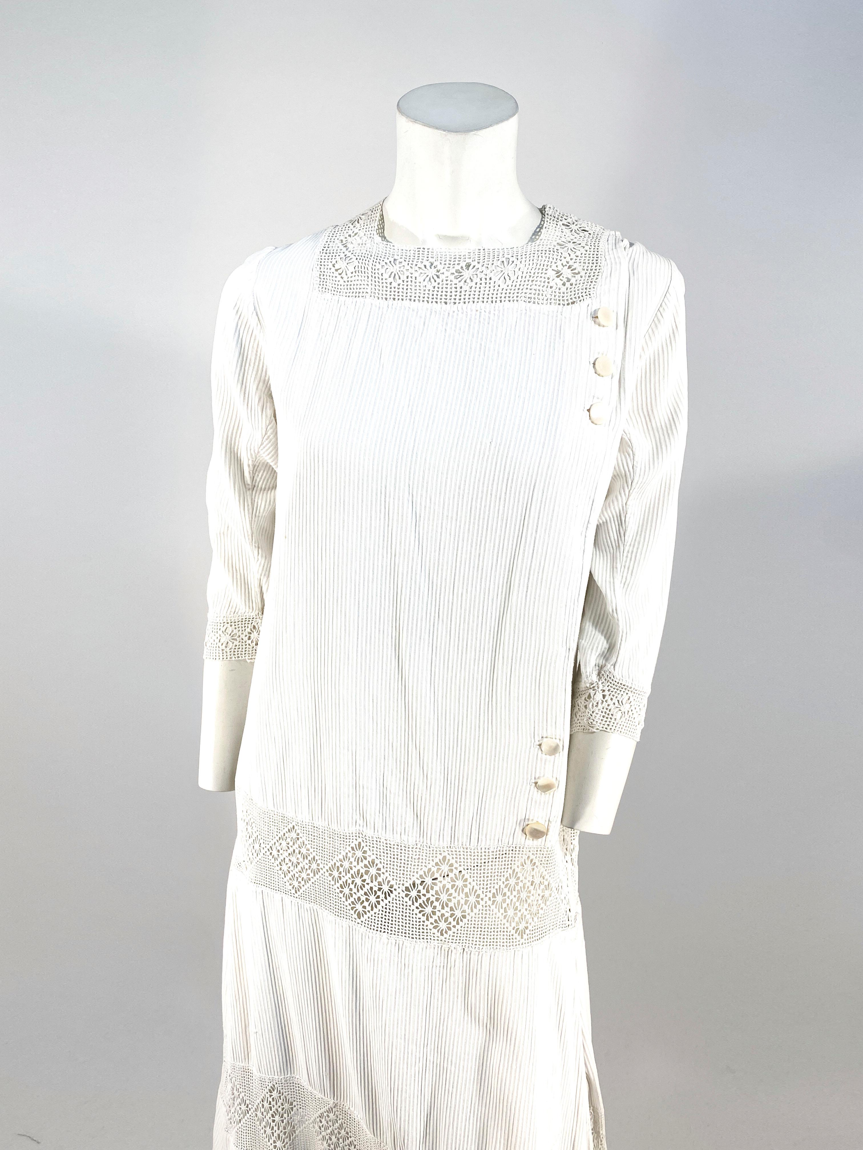 1920s white ribbed cotton day dress featuring a dropped waist band of hand crochet border that corresponds with the crochet on the elbow cuffs, skirt border, and square neckline. The asymmetrical front closure is finished with six seashell buttons.