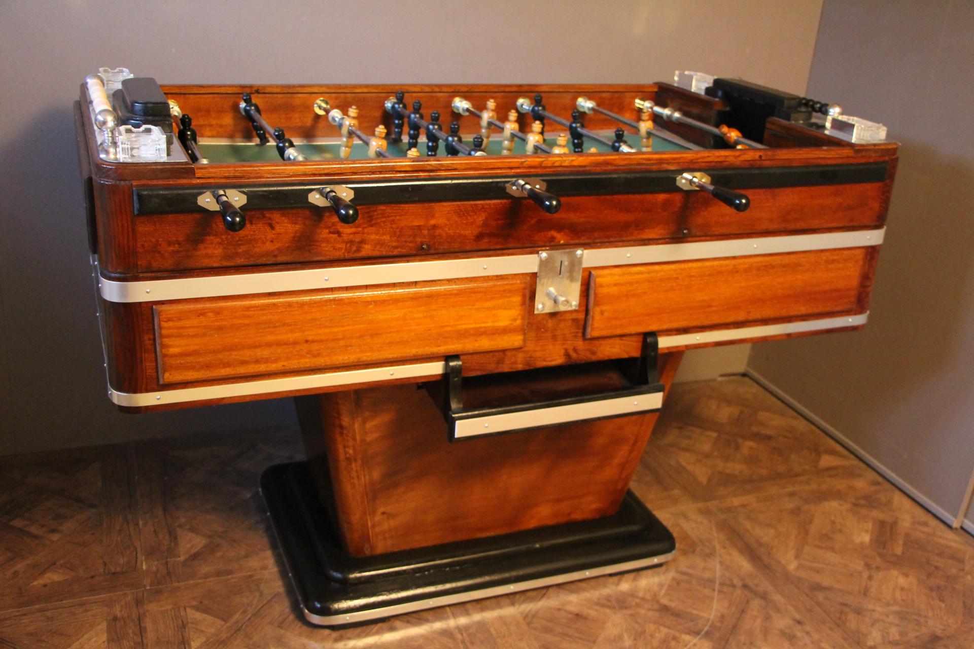 This foosball table features a honey color walnut. It still has got its four original ashtrays by Lalique, as well as all its polished aluminium pieces. Its players are made of turned wood and its field is green. It is coin operated.
Its shape is