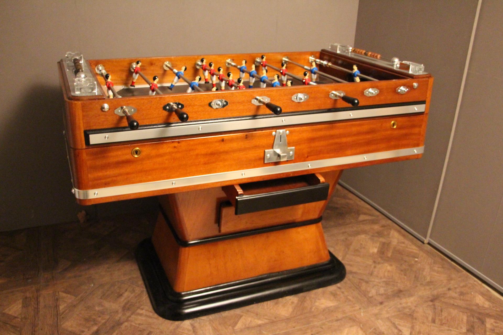 This foosball table is very unusual because it features a honey color wood. It still has got its four original ashtrays, as well as all its polished aluminum pieces. Its players are red and blue and its playground is black. Its shape is very elegant