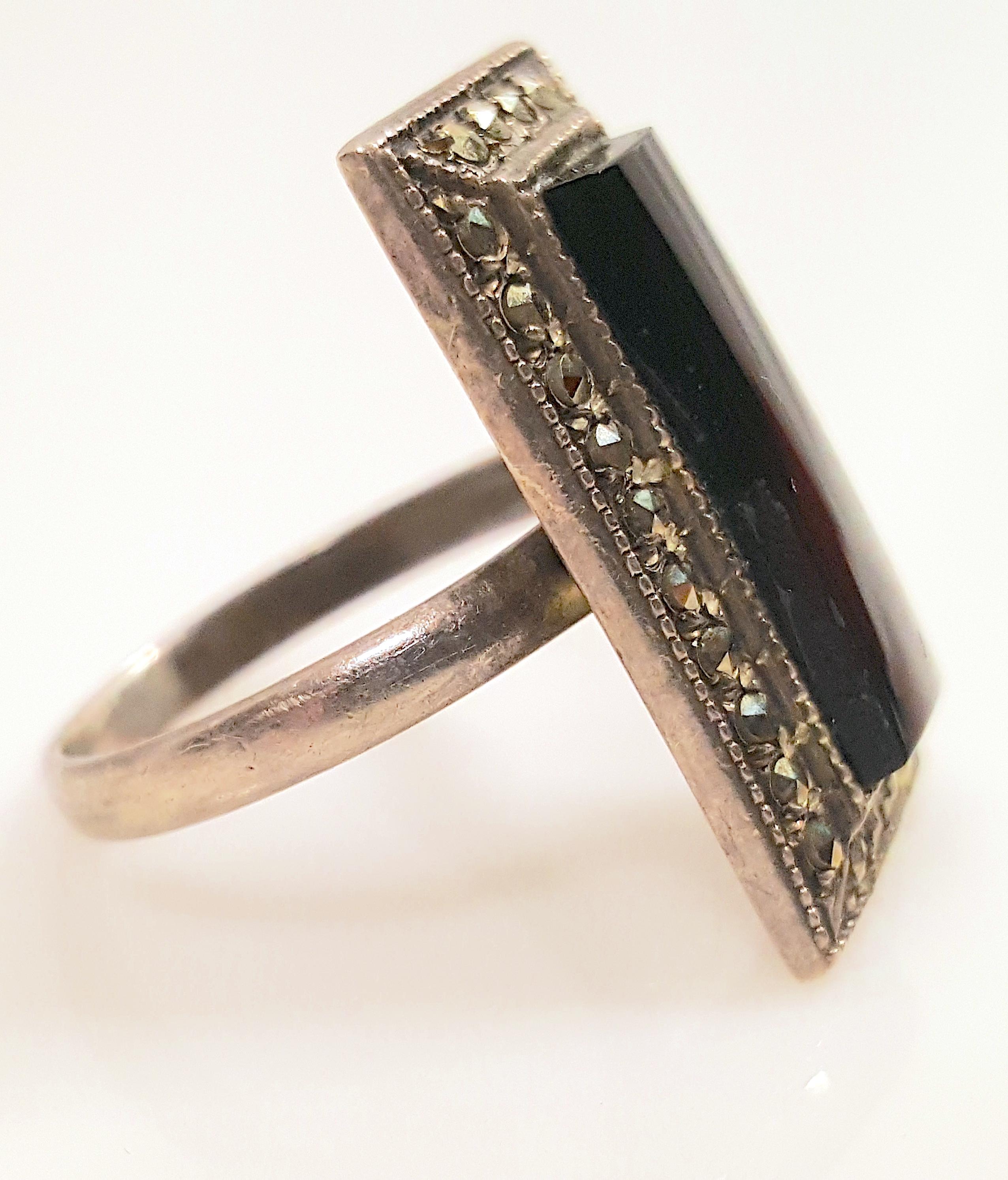 On this antique early Art Deco-period solitaire cocktail ring, a long rectangular mirror-cut black onyx is surrounded by 28 faceted marcasite stones. The American silver ring, whose face is 1 x 0.5 inches, is marked 
