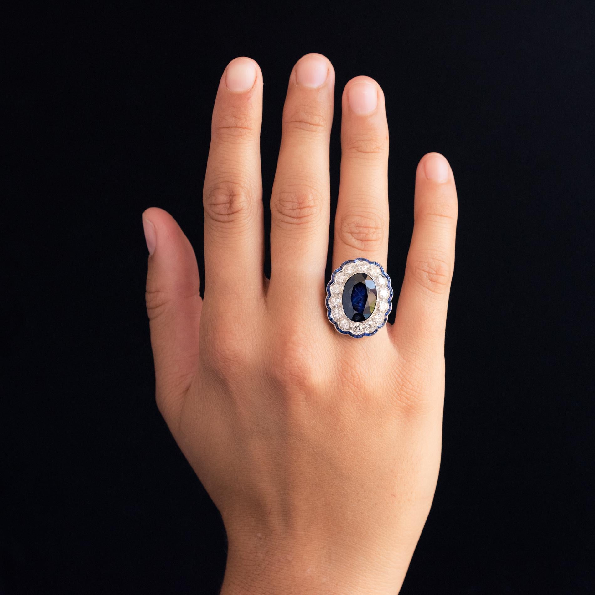 Ring in platinum.
Oval in shape, this magnificent Art Deco ring is adorned on its top with a blue sapphire supported in a millegrain setting, surrounded by antique brilliant-cut diamonds, bordered by calibrated blue sapphires on a multi-lobed