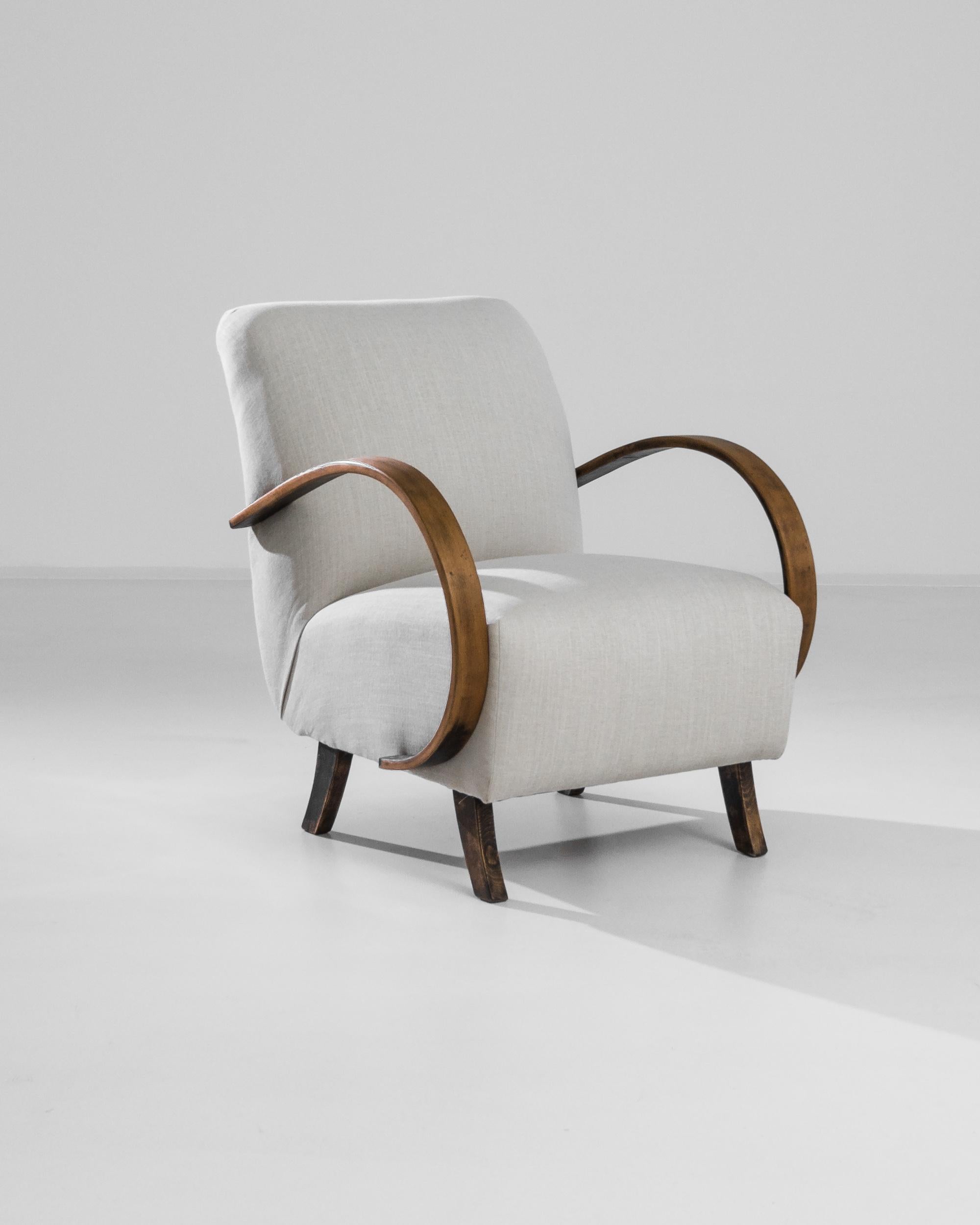 An armchair by Czech furniture designer Jindrich Halabala. Influenced by Modern and Art Deco design, a deep cushioned seat leans upon angled wooden feet, framed by swooping bentwood armrests. The smooth polish of the wooden arms, weathered to a rich