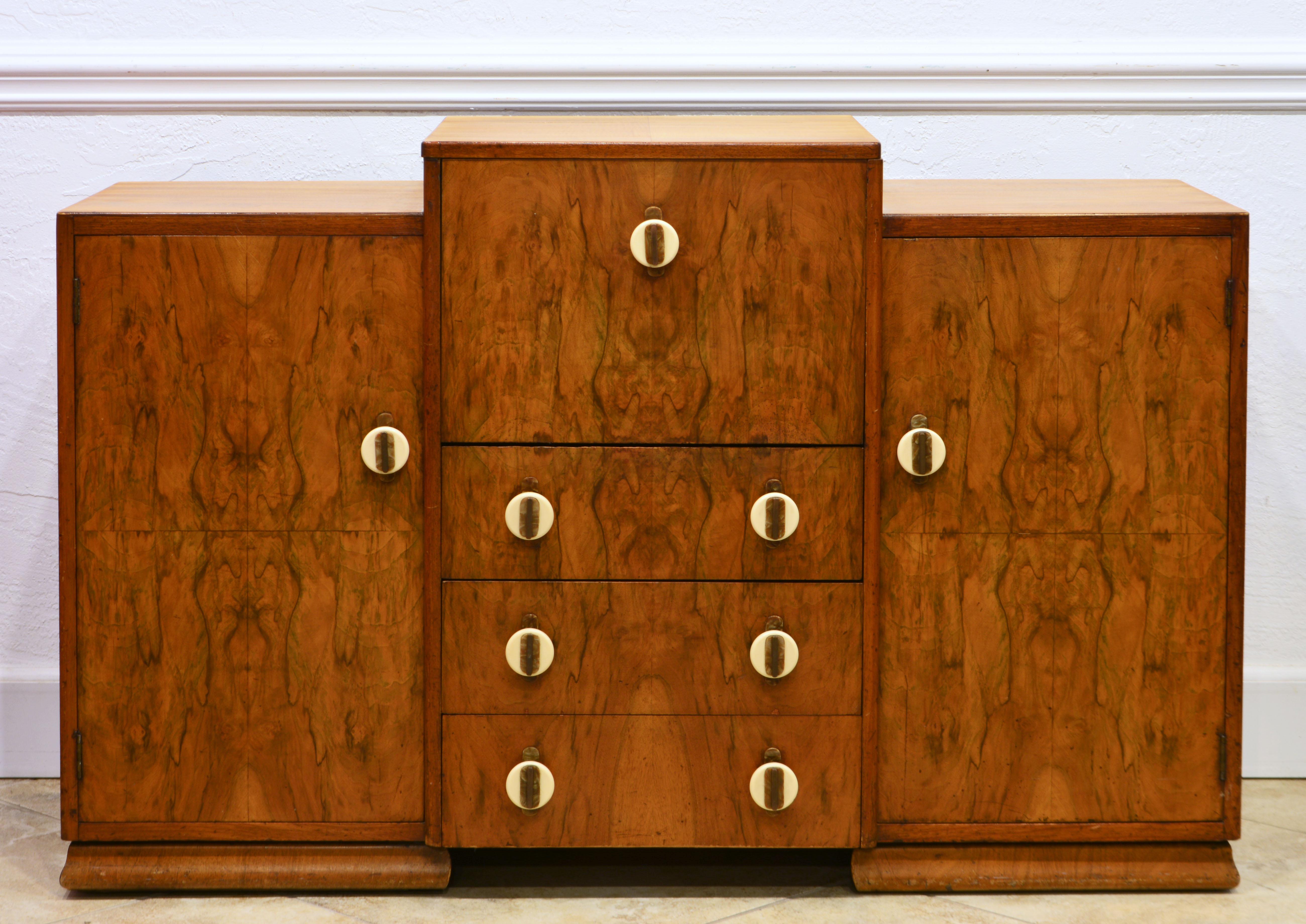 This Art Deco and Bauhaus style cocktail cabinet or bar dates to the 1930's and is likely made in England. It features a higher center section with three drawers under a mirrored and lit mixing unit which open top and door simultaneously. The center