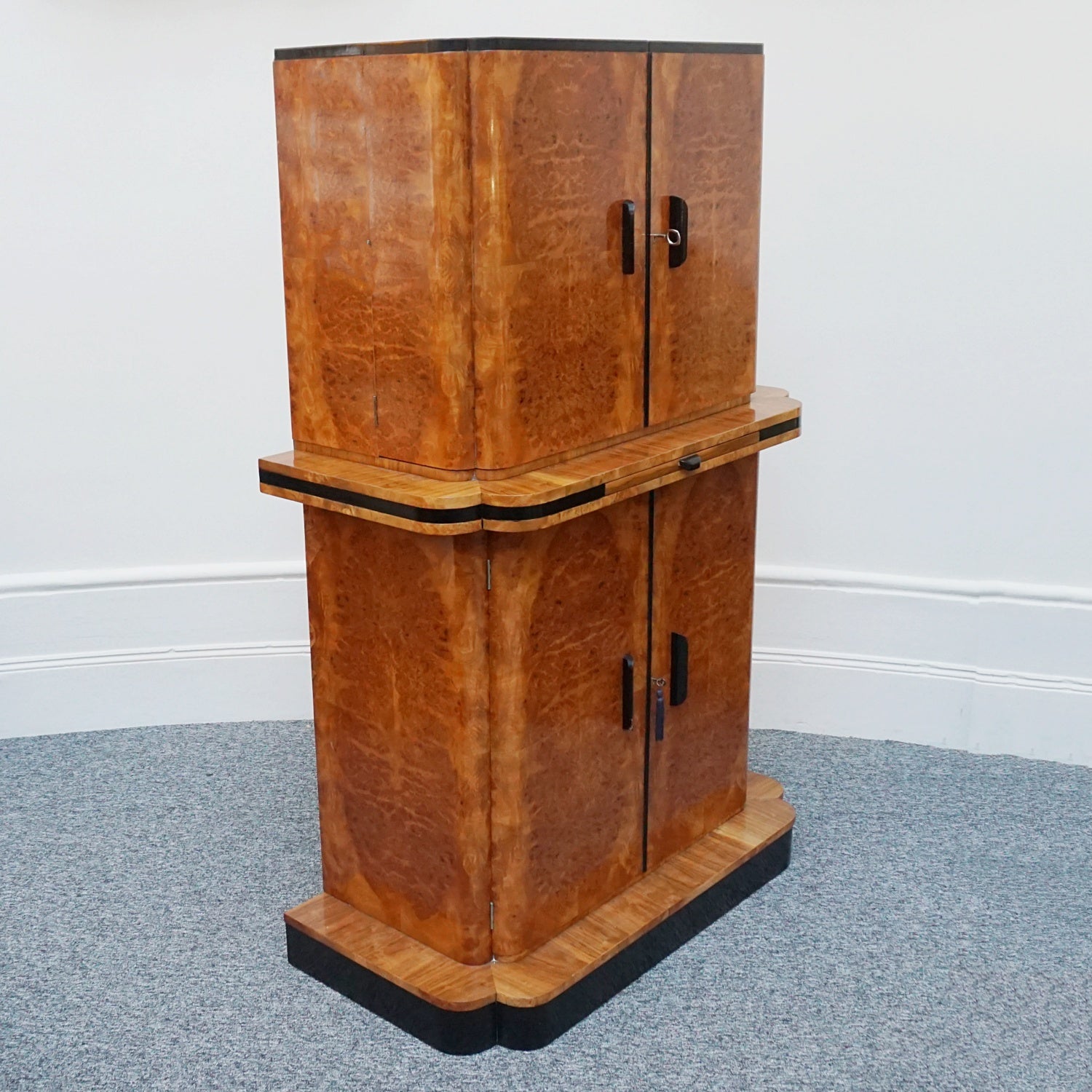 An Art Deco cocktail cabinet by Harry & Lou Epstein. Burr and figured walnut veneered with ebonised banding. Satin birch veneered upper mirrored and lit interior with original decorated mirrored glass. Pull out mirrored drinks tray to centre with