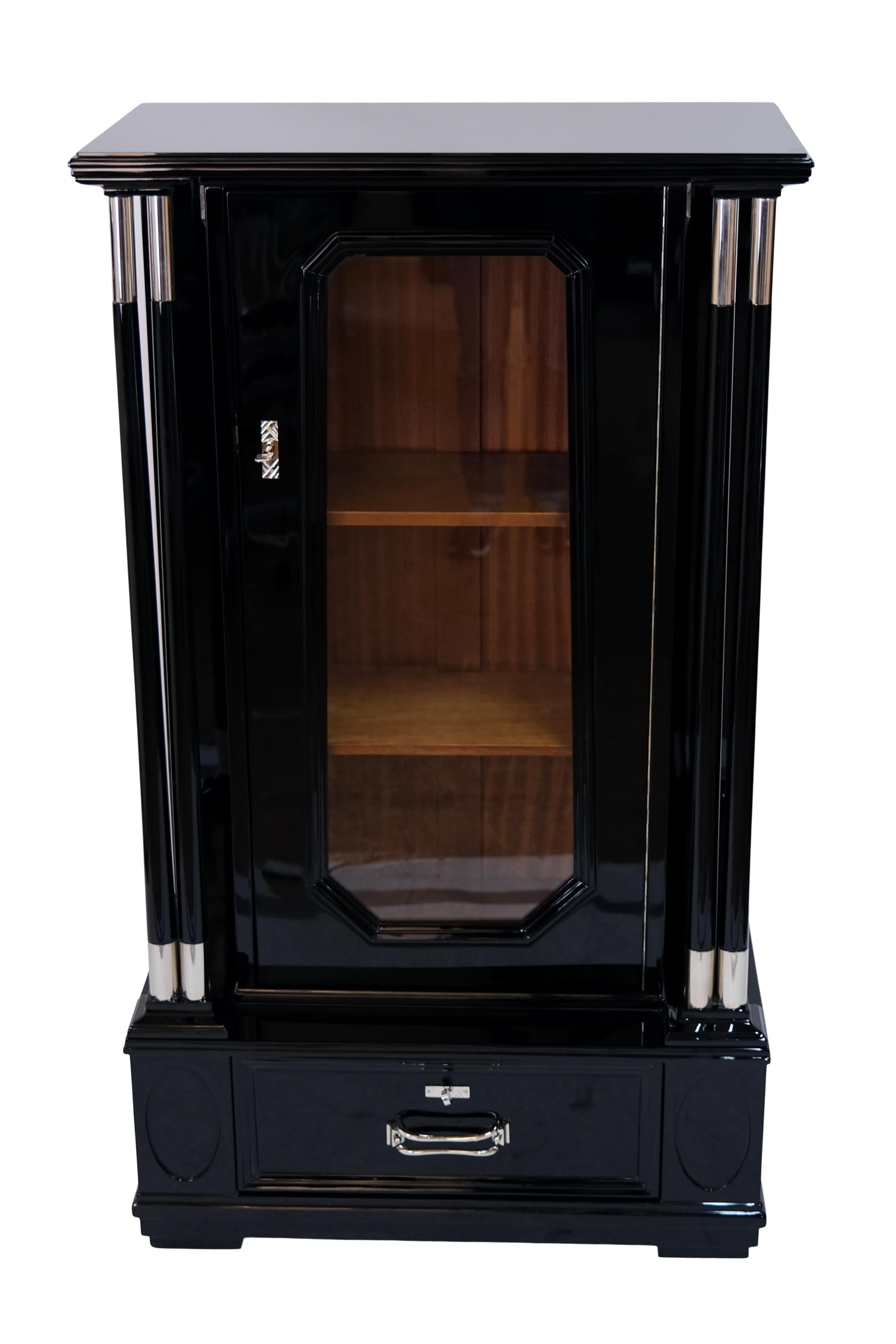 Furniture with columns, a central door and a drawer in the base area
Black piano lacquer with high gloss finish 
Nickeled details 
Glass in the front door 
Art Deco Patterns on the side

Austria, 1930s

Dimensions:
Width: 82 cm
height: 144