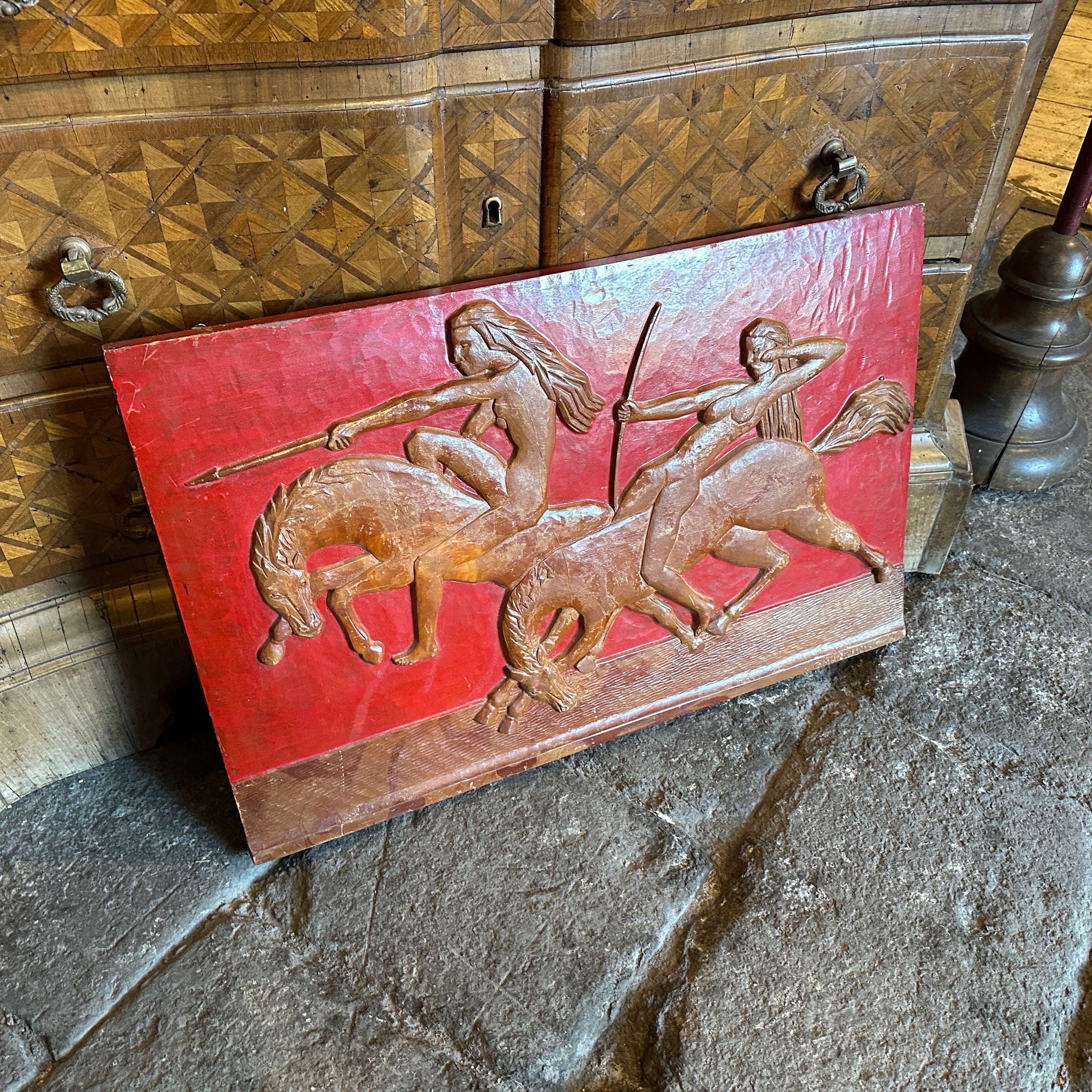 Ein erstaunliches handgeschnitztes Flachrelief aus Holz, das zwei Amazonen auf rotem Hintergrund darstellt. Es wurde von C. Mannini in den dreißiger Jahren realisiert, es ist auf einer Seite signiert. Es handelt sich um ein hervorragendes