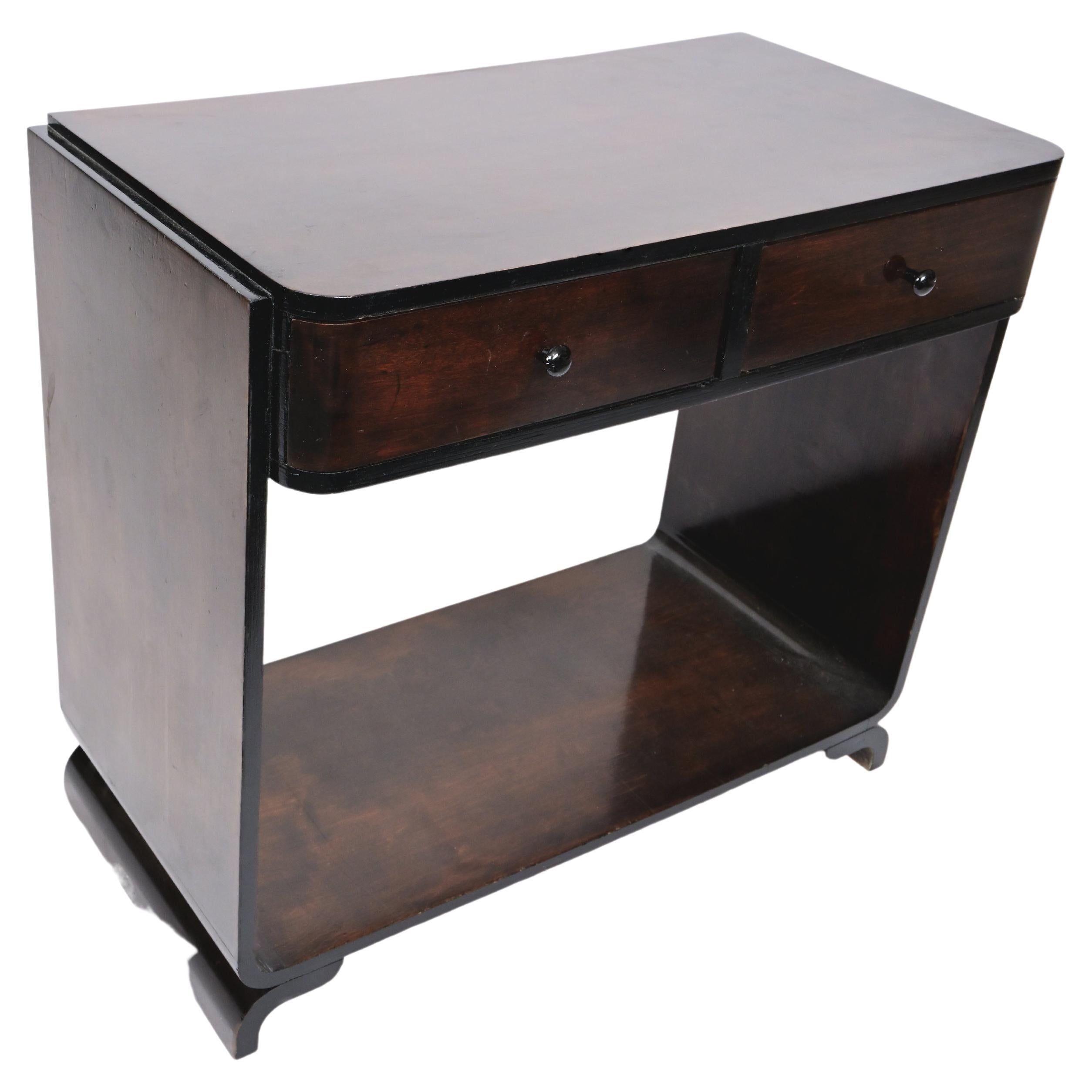 A black stained birch two tiered Art Deco side table featuring a curved bottom with two tapered and extended legs. Contains two drawers on the upper half. Circa 1930.