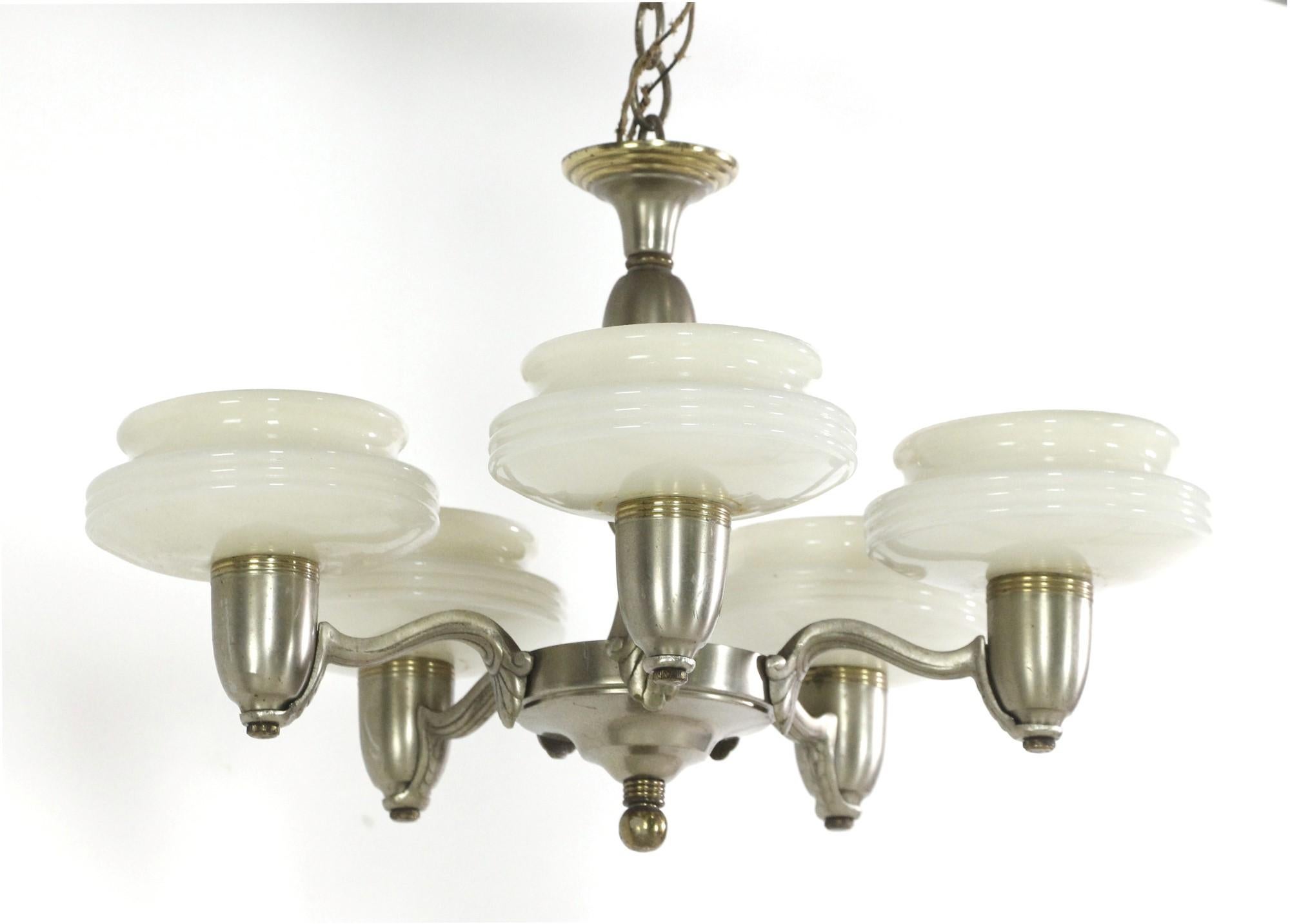 This Art Deco brushed nickel over brass chandelier features five-arms each fitted with a white slip shade. The arms each feature a leaf detail attaching it to the base of the light and the slip shades feature subtle lines. Cleaning and rewiring