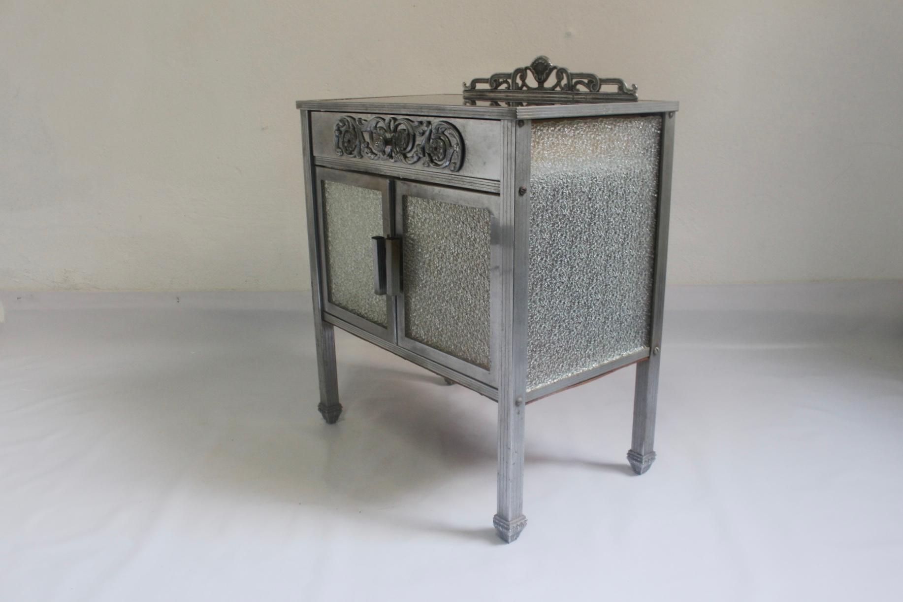 Art Deco cupboard from the 1930s, made from solid plated-nickel structure with smoke glass top and wooden shelf with four original glass panels. Drawer is made of wood and embellished with carved metalwork.
Condition: Good vintage condition, with