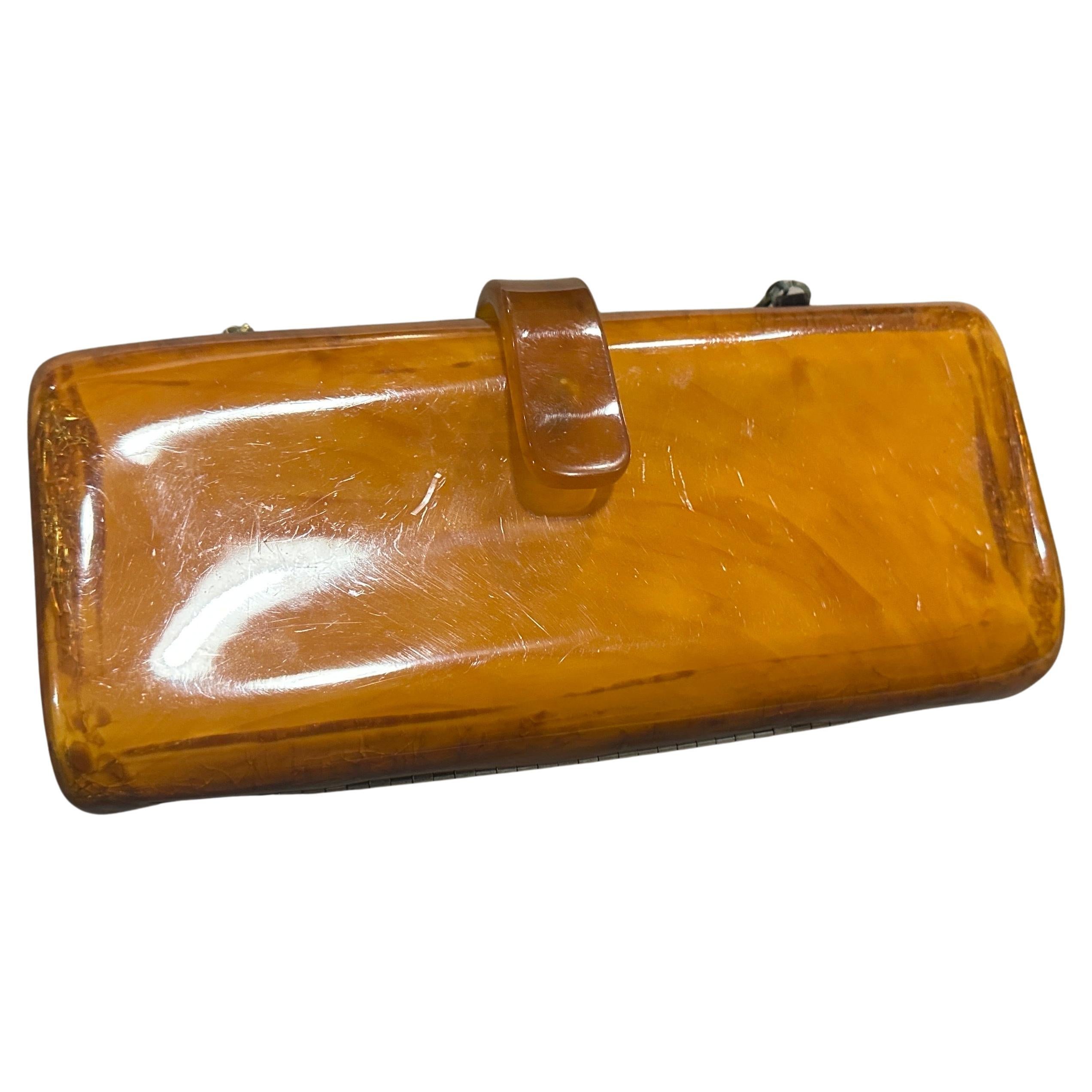 This 1930s Art Deco evening shoulder bag is crafted from rich amber-hued Bakelite, a highly collectible early plastic known for its smooth, glossy finish and distinctive marbling. The bag features a sleek, rectangular hard case design with softly
