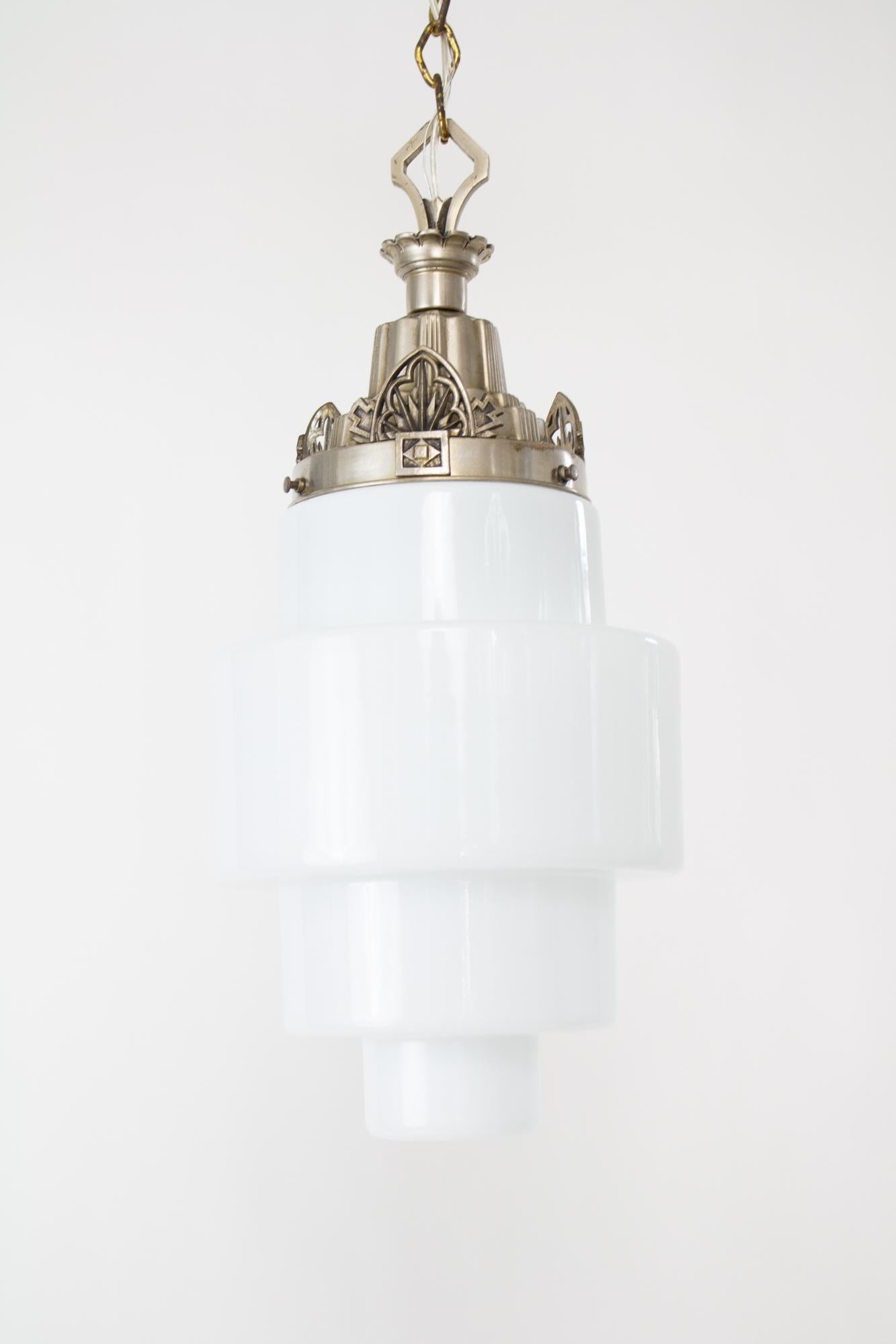 Art Deco milk glass pendant with a nickel plated cast metal fixture. The fixture has a long top loop and crown shaped glass holder with a geometric flare design. The canopy is a nickel/brass combination with a classical art deco geometric pattern.
