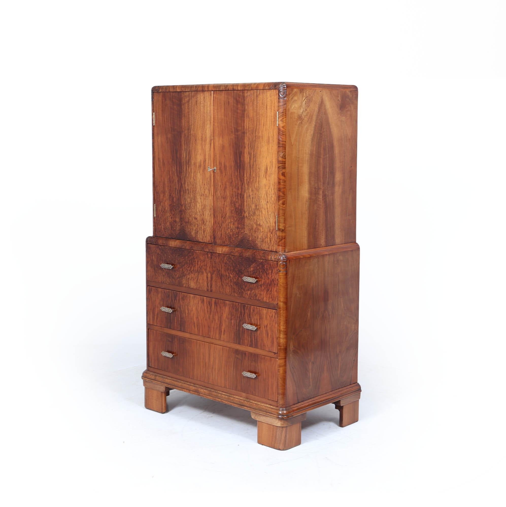 Art Deco Tallboy 
A tall and handsome English Art Deco cabinet in beautifully figured walnut, circa 1930s. The upper section features a two-door cupboard with original key, above a bank of three drawers with stepped chrome handles. Subtle carved