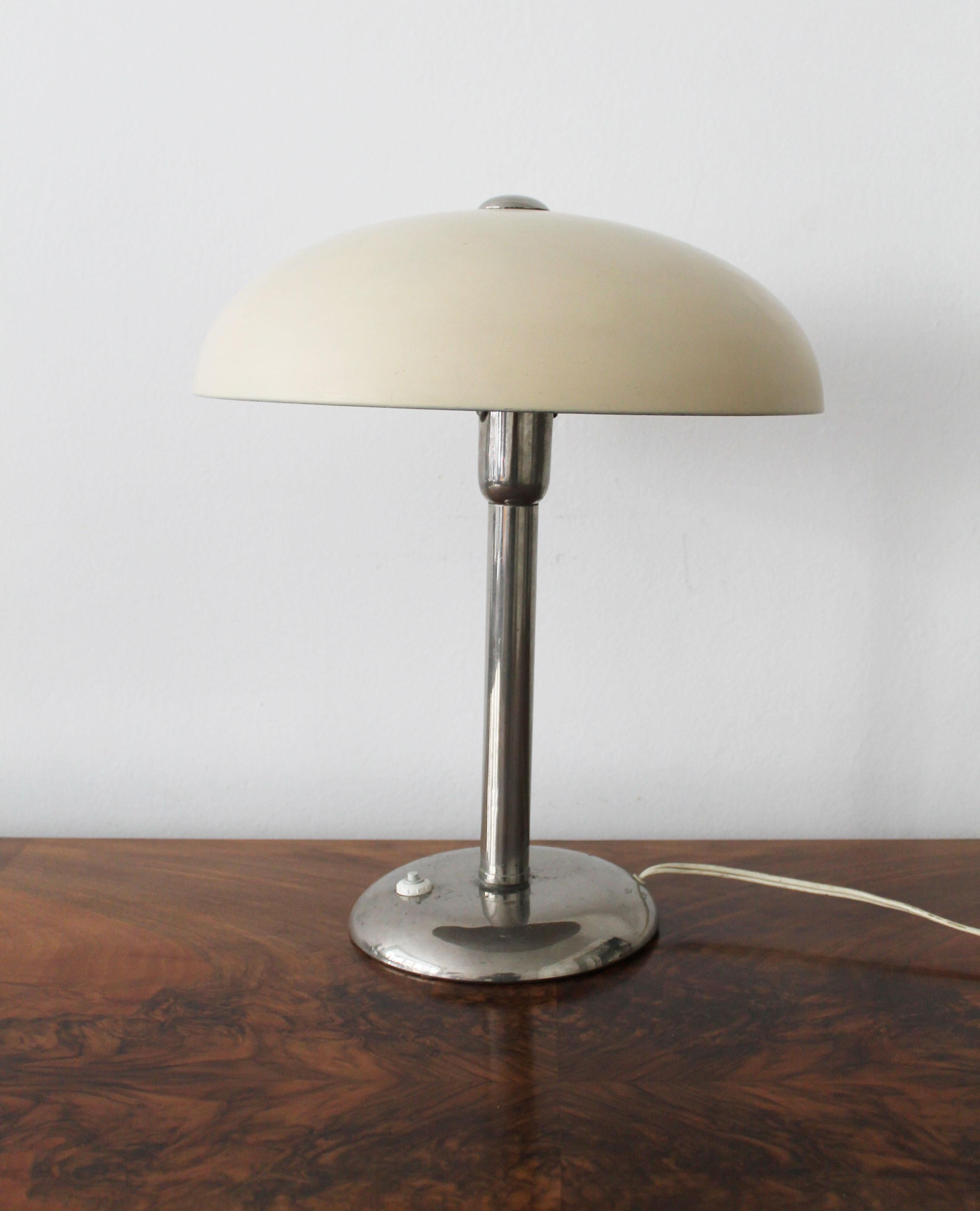 This 1930’s Modernist table lamp is made of chromed steel with a lampshade painted in a cream colour. The bulb is positioned vertically, right underneath the large shaped mushroom-like shade that is fixed to the base by a single flat screw on the