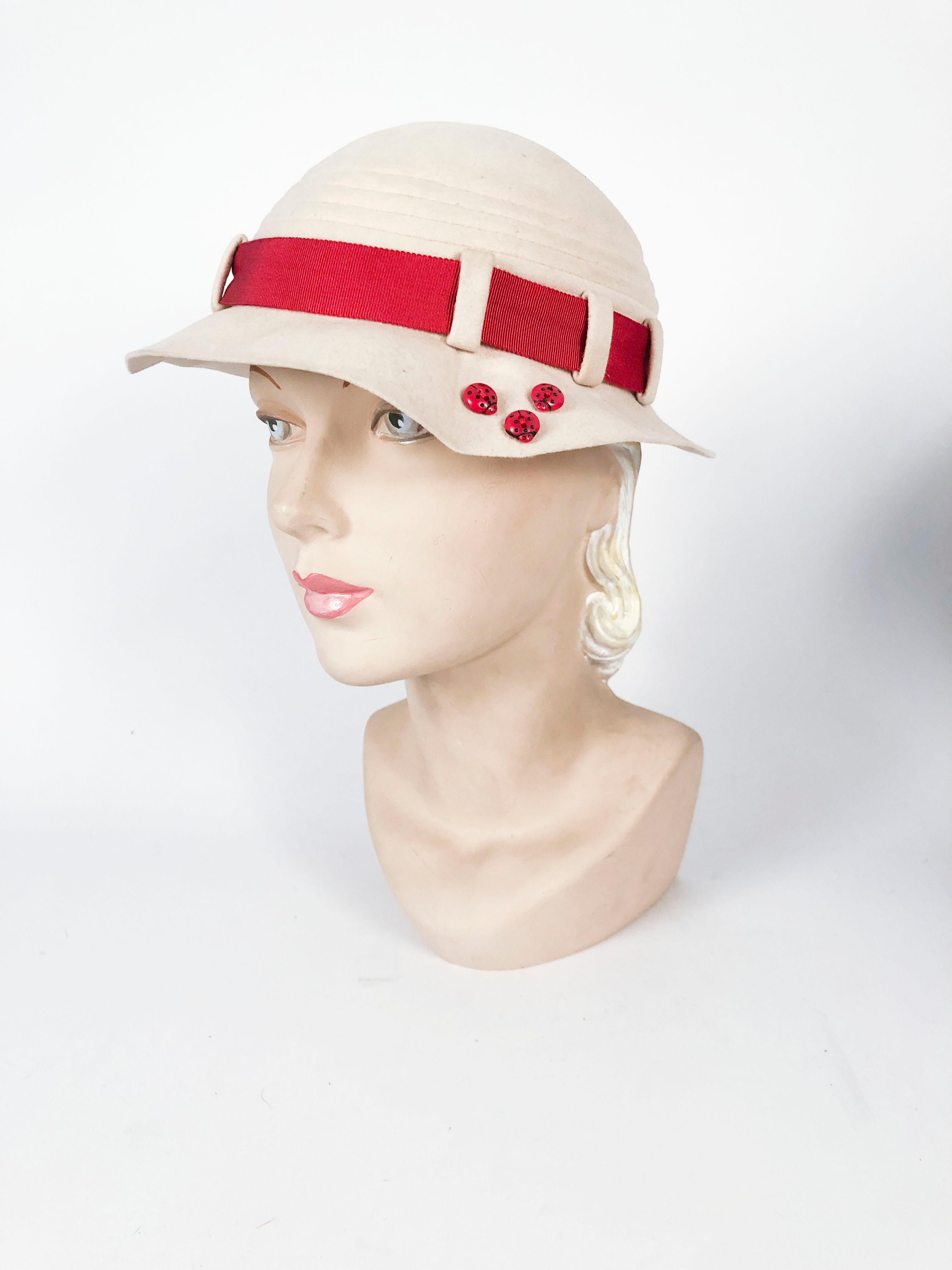 Geformter beiger Filzhut aus den 1930er Jahren mit rostfarbenem Ripsband und Art-Déco-Knopf. Die geformte Krempe hat 3 Marienkäfer-Akzente, die dem Hut eine asymmetrische Note verleihen. 