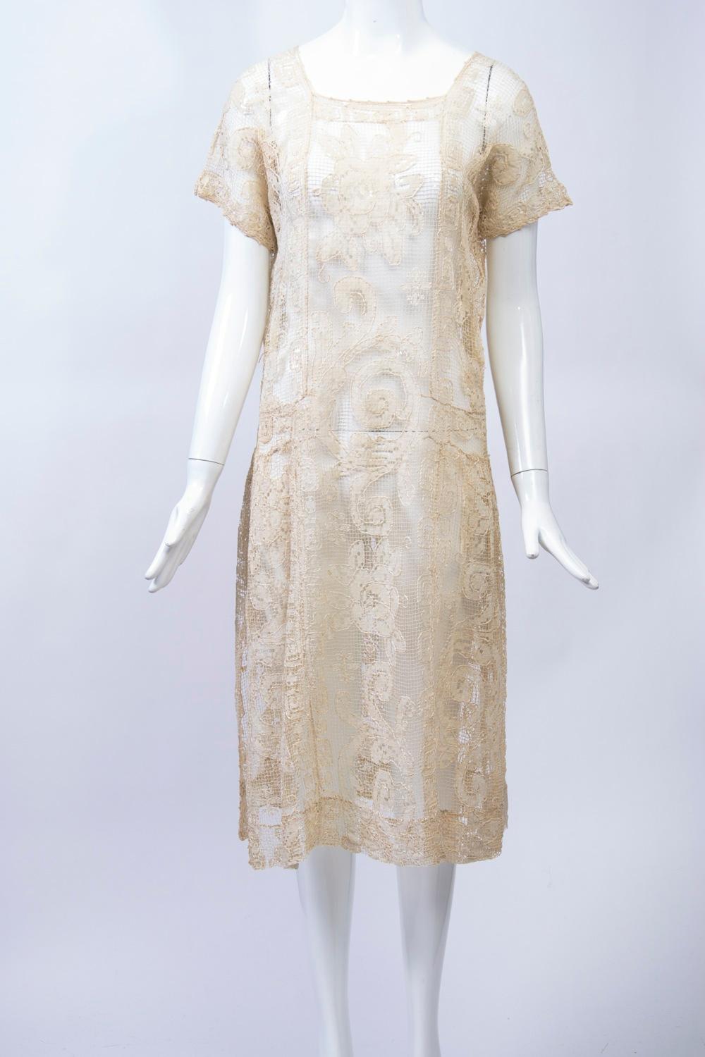 1930s beige lace dress featuring a square neckline, short sleeves, and a dropped waistline with slight shirring at the sides, where there are slits at the hem; the front and back panels extend straight from the neckline to the calf-length hem. Add a