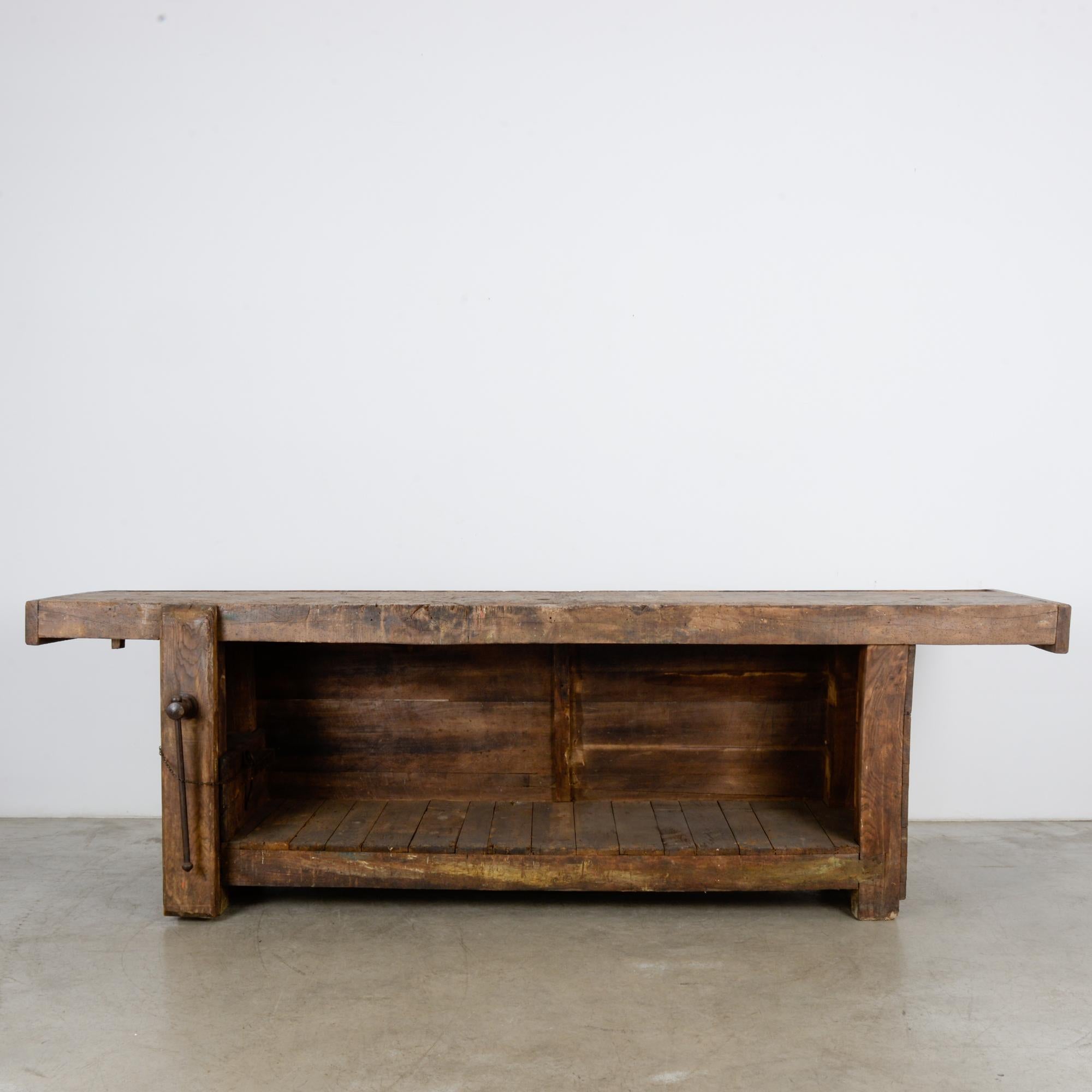 A wooden work table from Belgium, circa 1930. A rugged, dependable construction, composed of a sturdy worktop astride a trapezoid bench, mounted with a vertical wooden vise. A sunken channel runs along the length of the worktop, creating a sanctuary