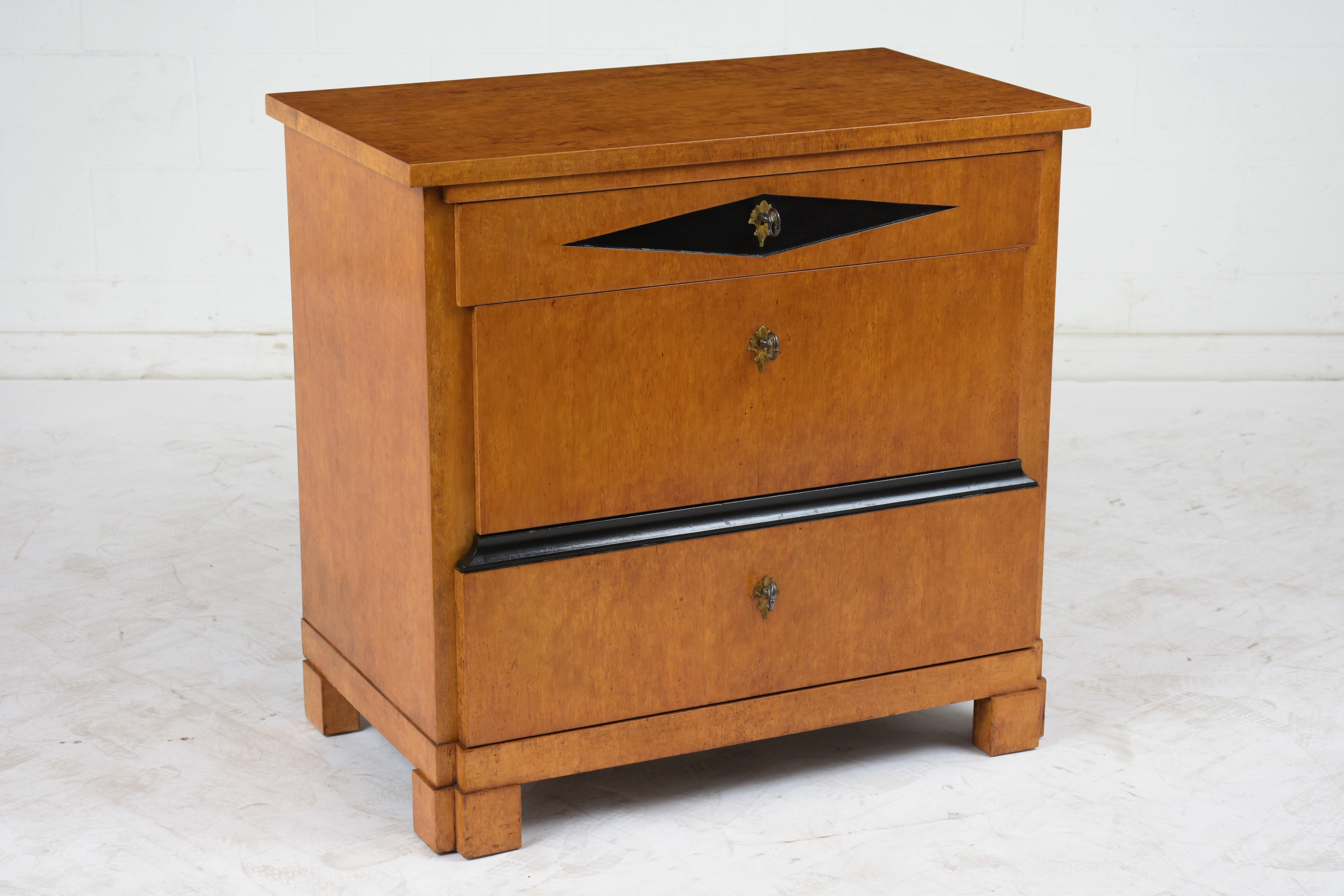 This 1930s Biedermeier-style chest of drawers is covered in birch wood veneers and finished in a light walnut color with black accents. There are three drawers with carved notches for opening and keyhole plates. The drawers are lined in silk with