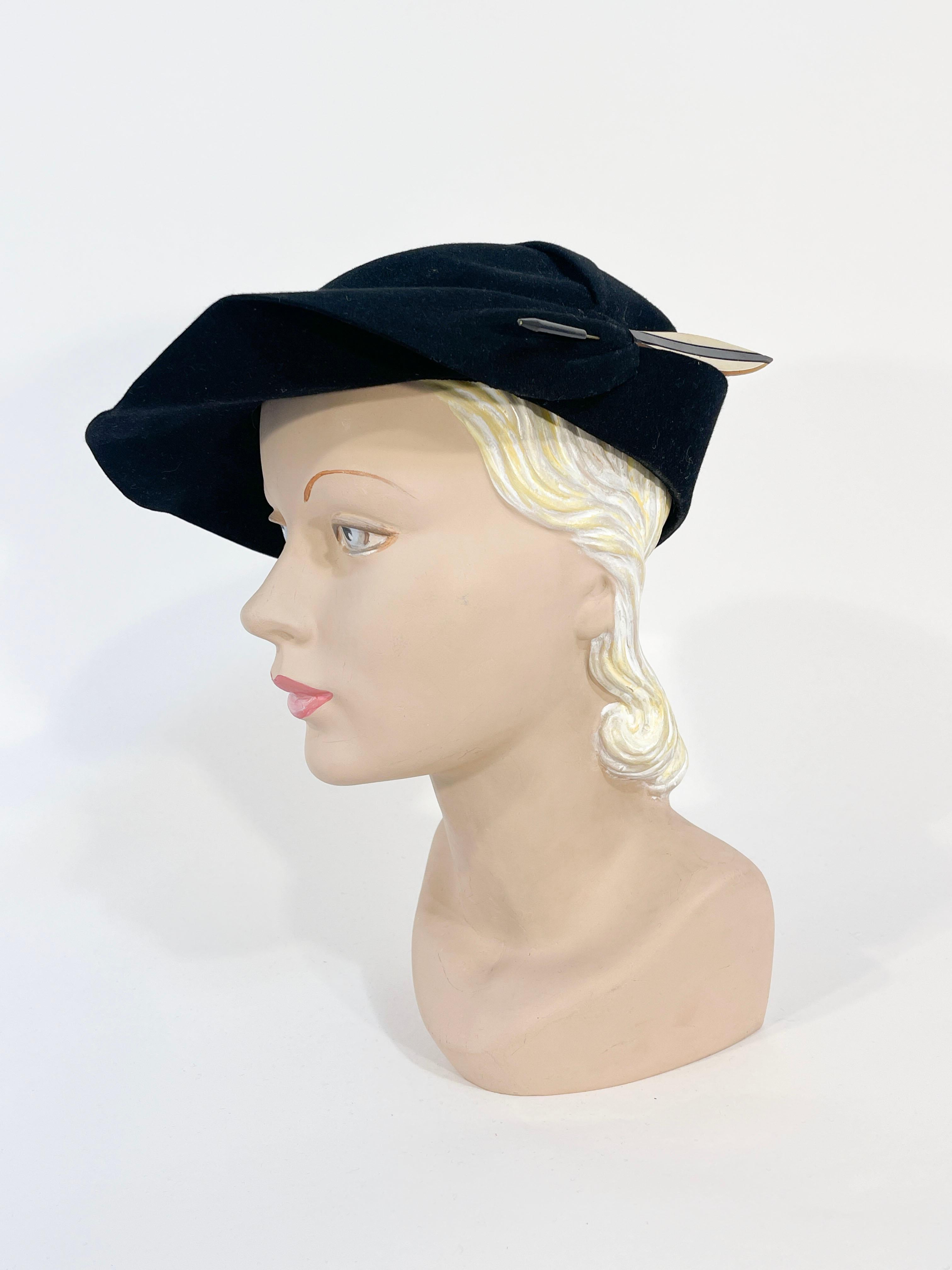 Chapeau asymétrique des années 1930 en laine feutrée noire, avec un bord en forme de 2,5 pouces de large, une couronne arrondie et plissée et une longue épingle à chapeau en celluloïd noir et blanc en forme de plume Art déco. La goupille est