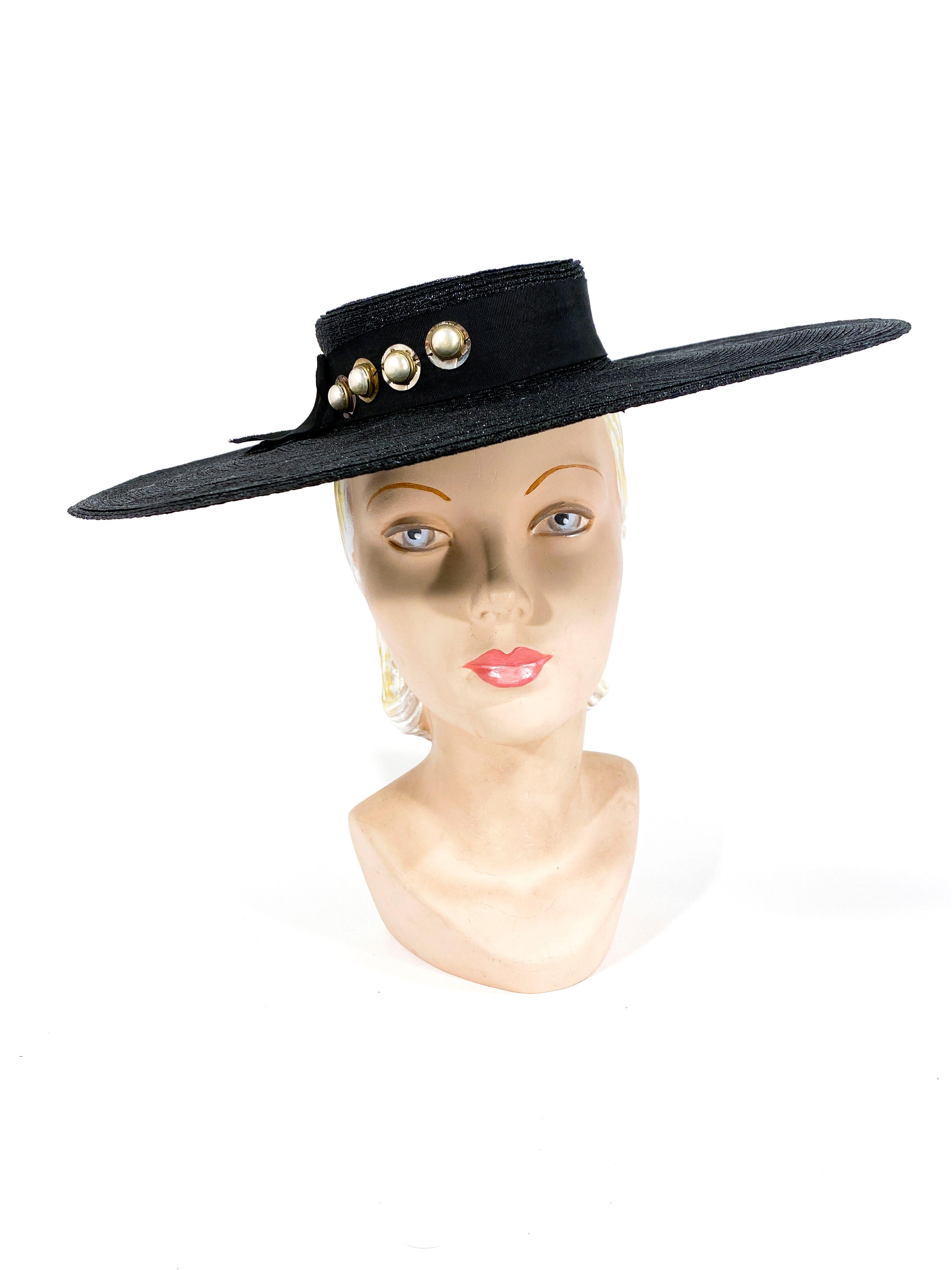 1930s black saucer hat made of a coated and woven straw. The wide-brim is wide, structured, and stiff. The shallow crown is hand sculptured and adorned with a grosgrain hat band. The band itself is decorated with large sequin and pearl buttons.
