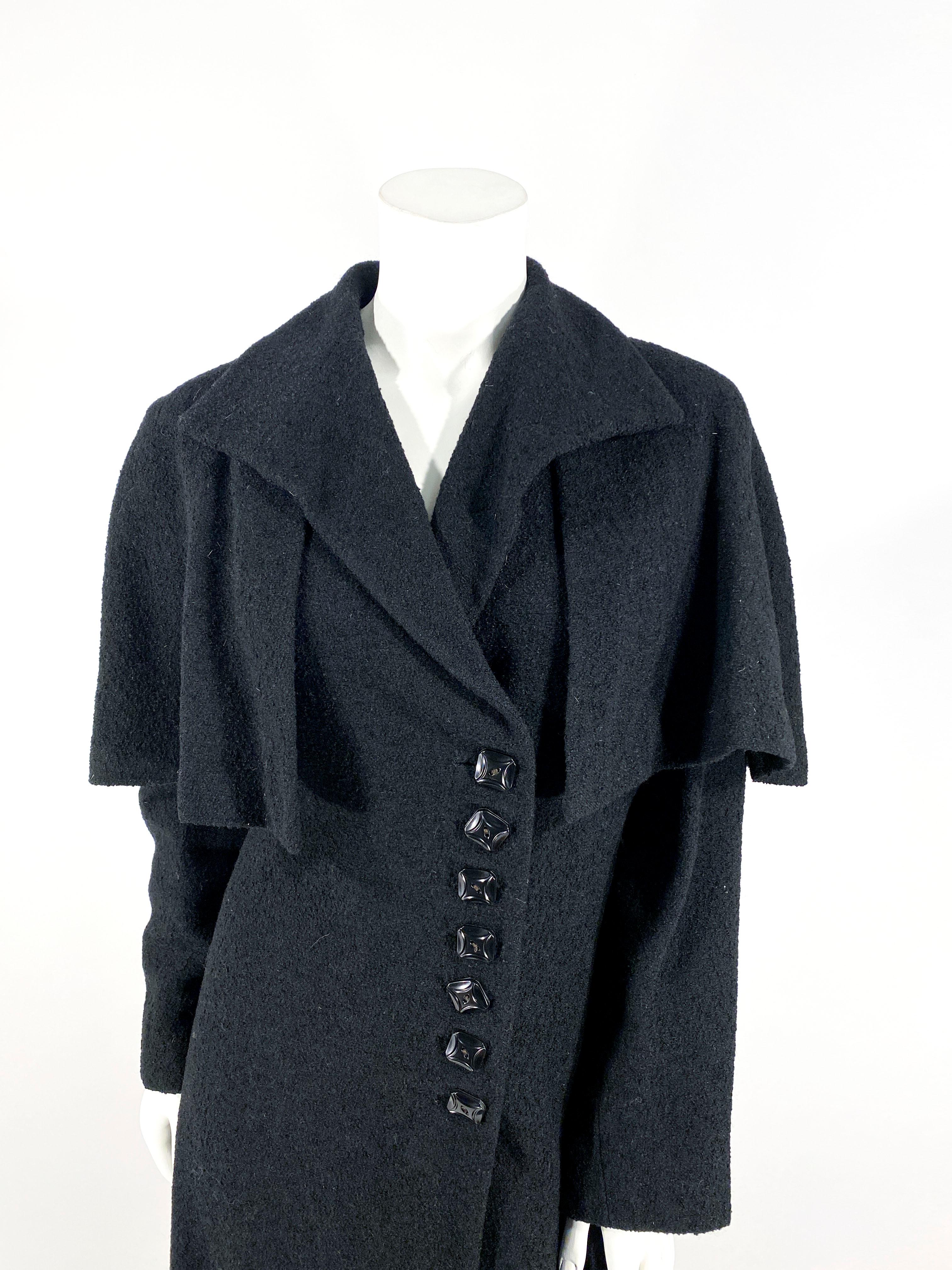 1930s black wool coat featuring an A-line silhouette, full length sleeves, a sting-ray collar and an attached capelet. The front closure has a series of black art deco buttons that match the rows of buttons on the back of each sleeve. The coat is