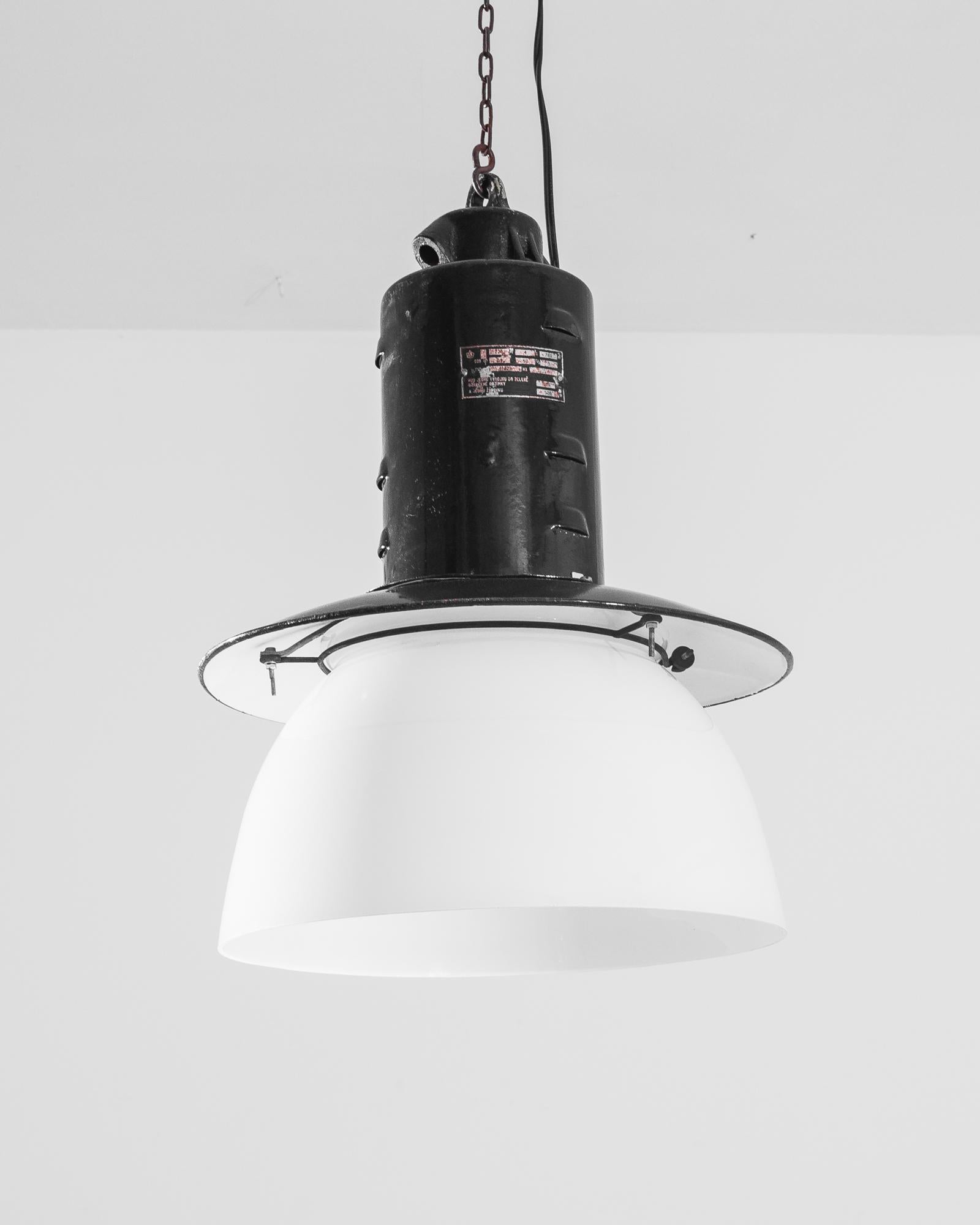 A metal industrial lamp from Czechia, produced circa 1930. Black metal cylinder suspended by chain from ceiling, blossoms into a shade covering a white glass bowl. This vintage lamp is an industrial stalactite from Central Europe stamped by the
