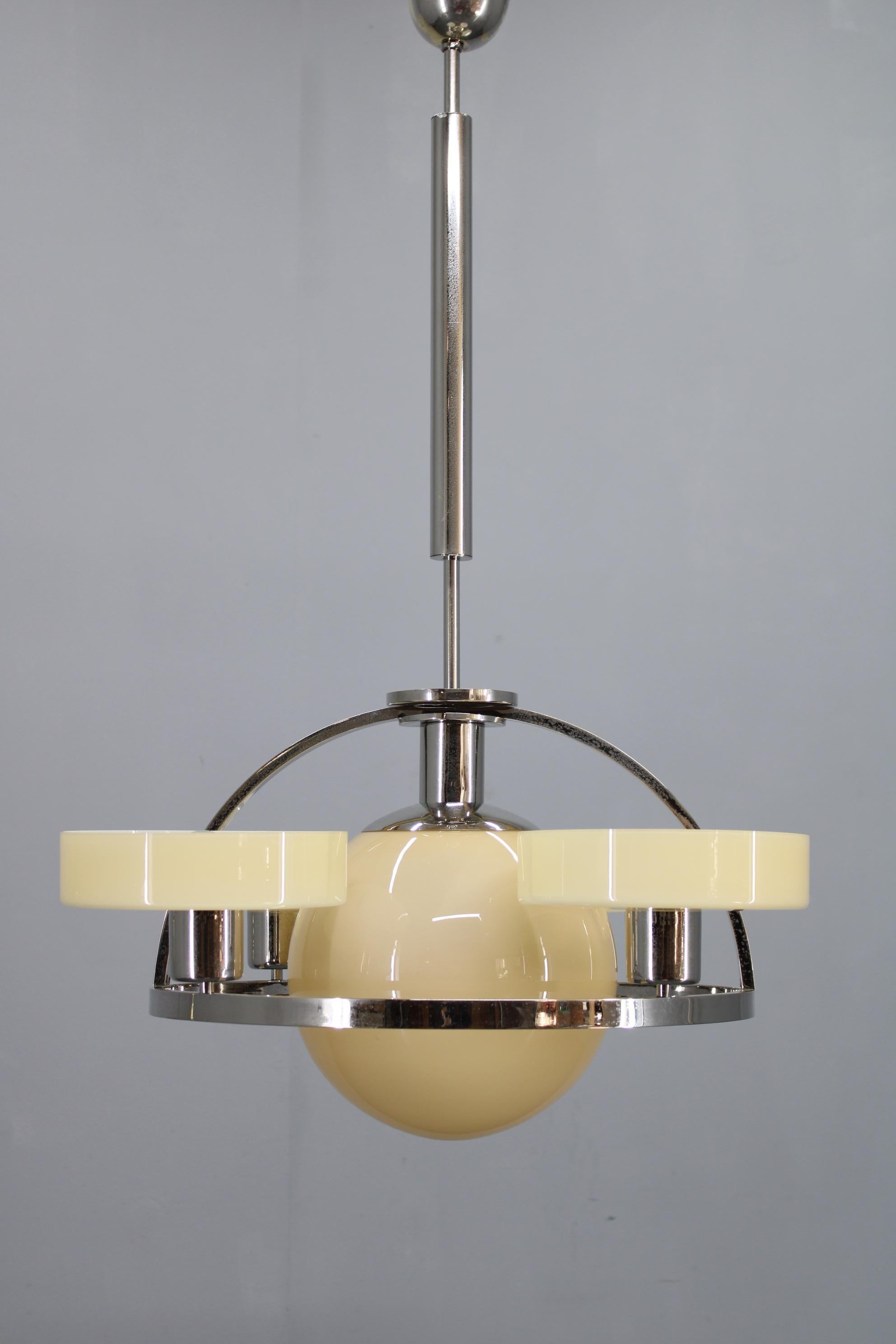 A sculptural Art Deco chandelier produced in Czechoslovakia during the 1930s, constructed with a polished chrome-plated metal body and five opaline beige–cream glass shades. The combination of chromed geometric arches and warm, softly diffusing