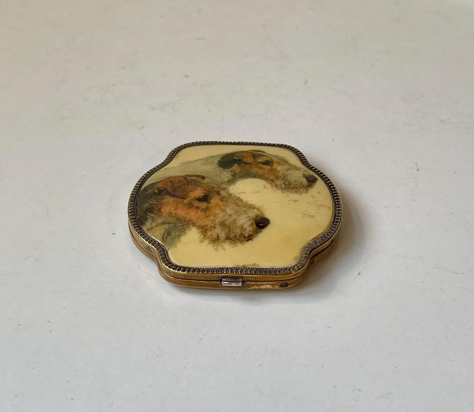 A rare cushion shaped powder compact featuring a pair of well-groomed fox terriers. Its exterior is executed in gold plate and enamel. The interior is intact with powder compartment and a small integrated circular mirror. It was made during the