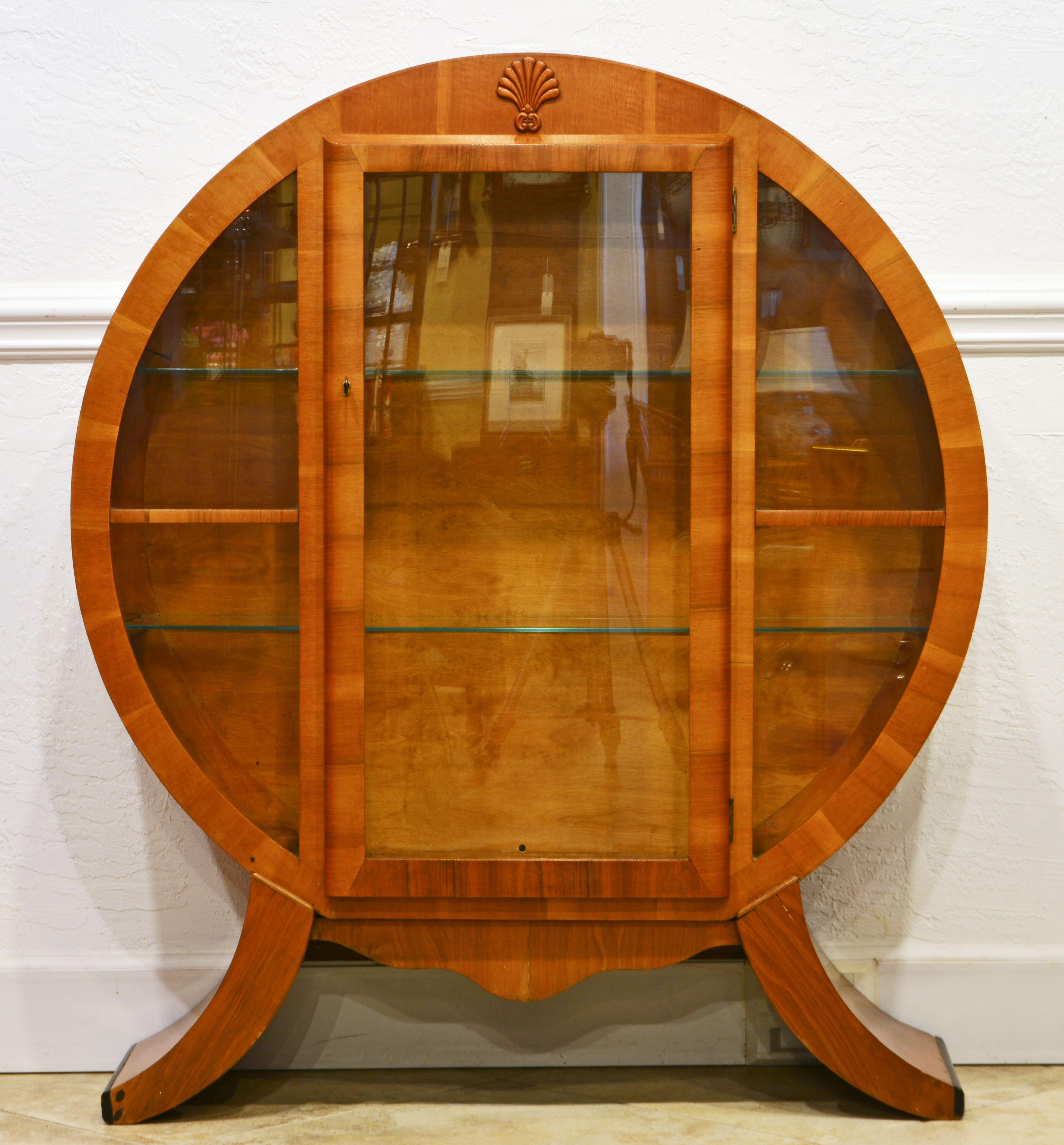 This spectacular English light walnut display cabinet features a circular body resting on elegantly curved feet with ebonized edges. The door opens up to an illuminated interior with two long glass shelves. The back of the interior is accented by