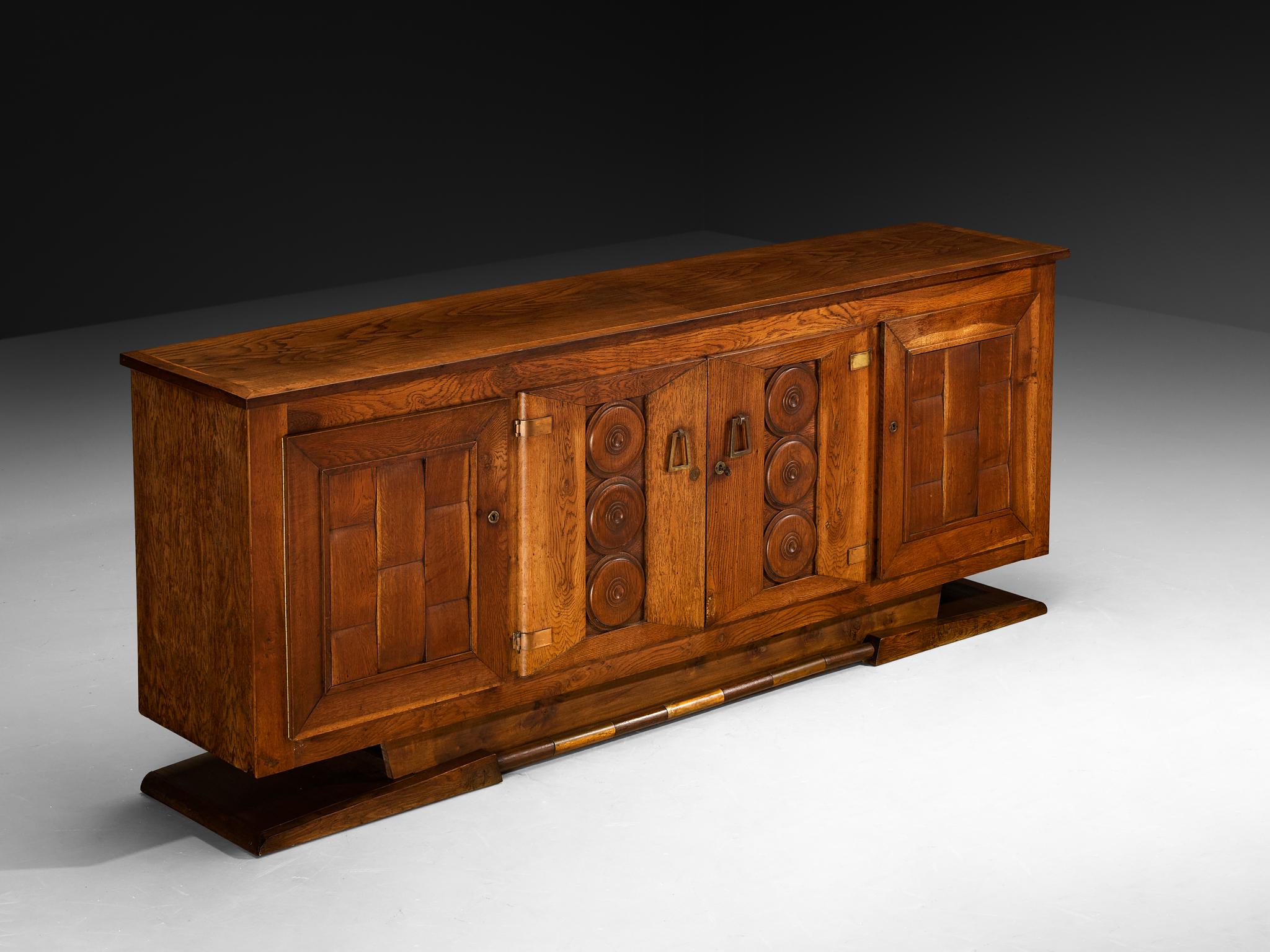 Buffet, chêne, laiton, France, années 1930 

Ce buffet est une belle expression du design Art déco français des années 1930. La silhouette générale est horizontalement emphatique, un bloc architectural au sol reposant sur un socle trapézoïdal qui