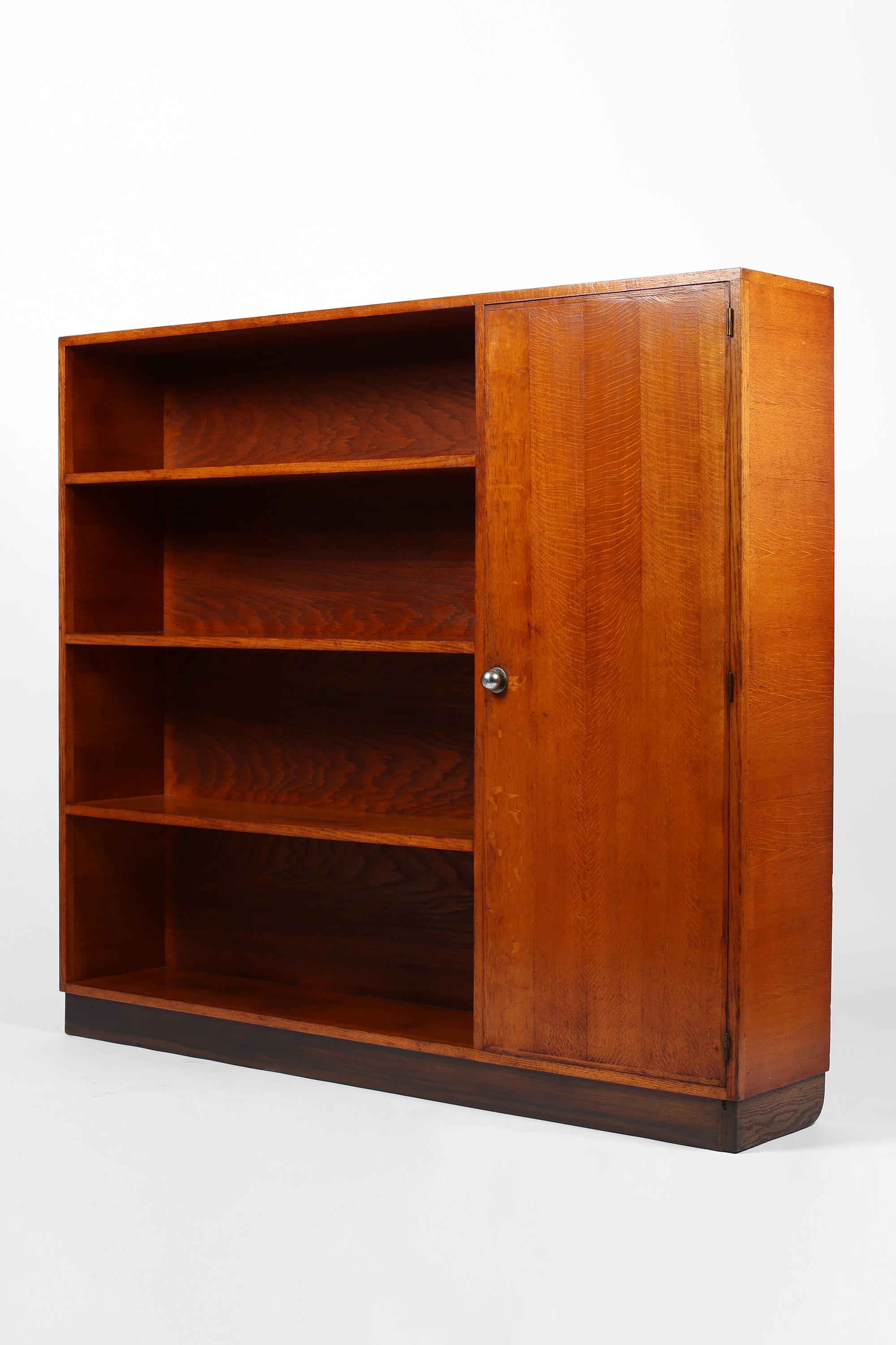 A slim modernist Art Deco bookcase constructed from richly patinated oak. Featuring four open shelves and single cupboard compartment, sitting on a recessed blackened plinth base. The cupboard retains its original chromed bronze door pull and opens