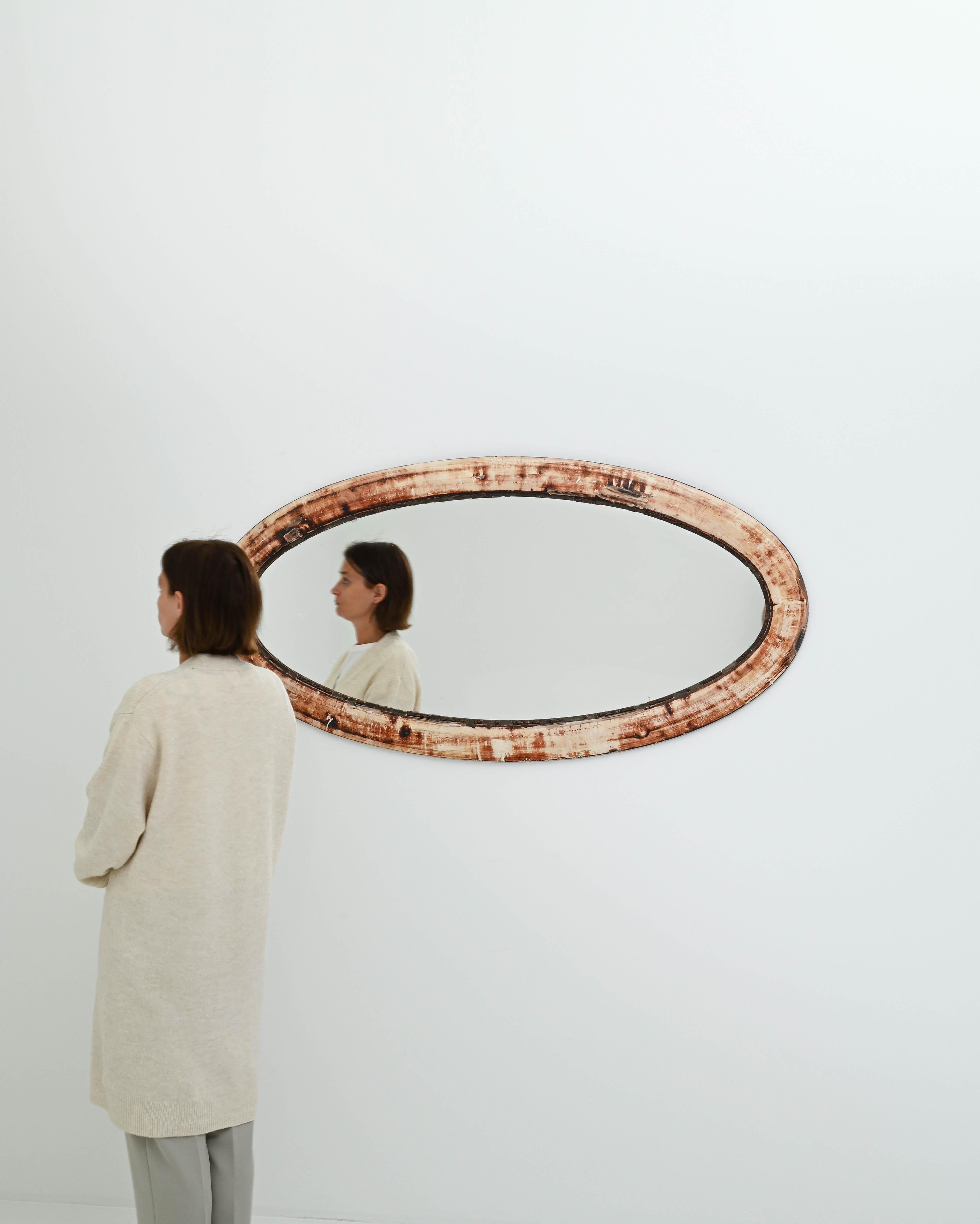 A wooden mirror created in 1930s France. A heavy patina and oval shape give this wall mirror a mysterious presence. Through time the wooden frame has developed a fiery patina, consisting of perplexing and eye-pleasing beiges, maroons, and reds.