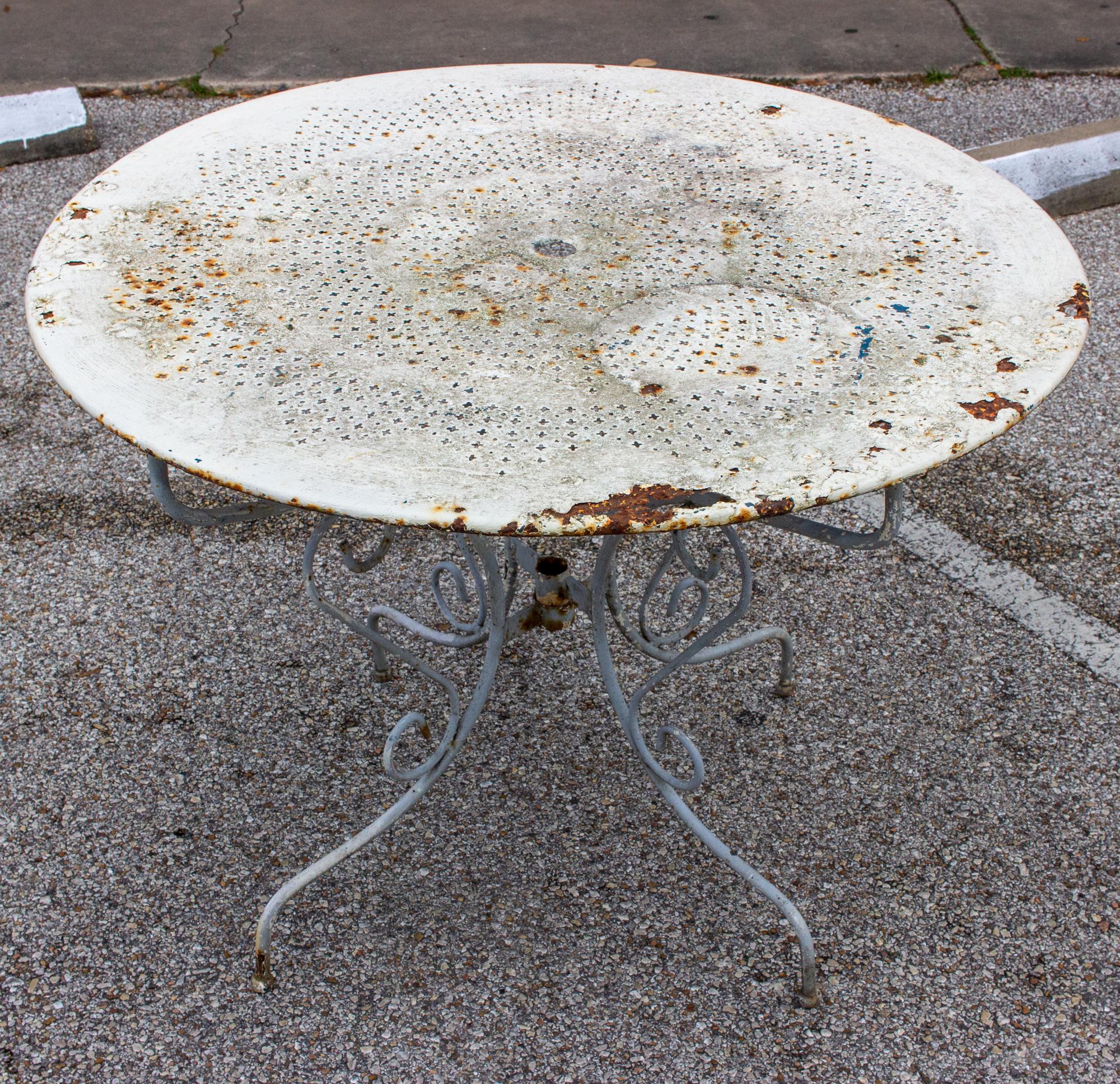 This is a rustic painted metal garden table we sourced in northern France. The base is painted a pale gray color with some white and blue paint peeking through (likely from previous paint jobs). The top is painted in white. The center of the top