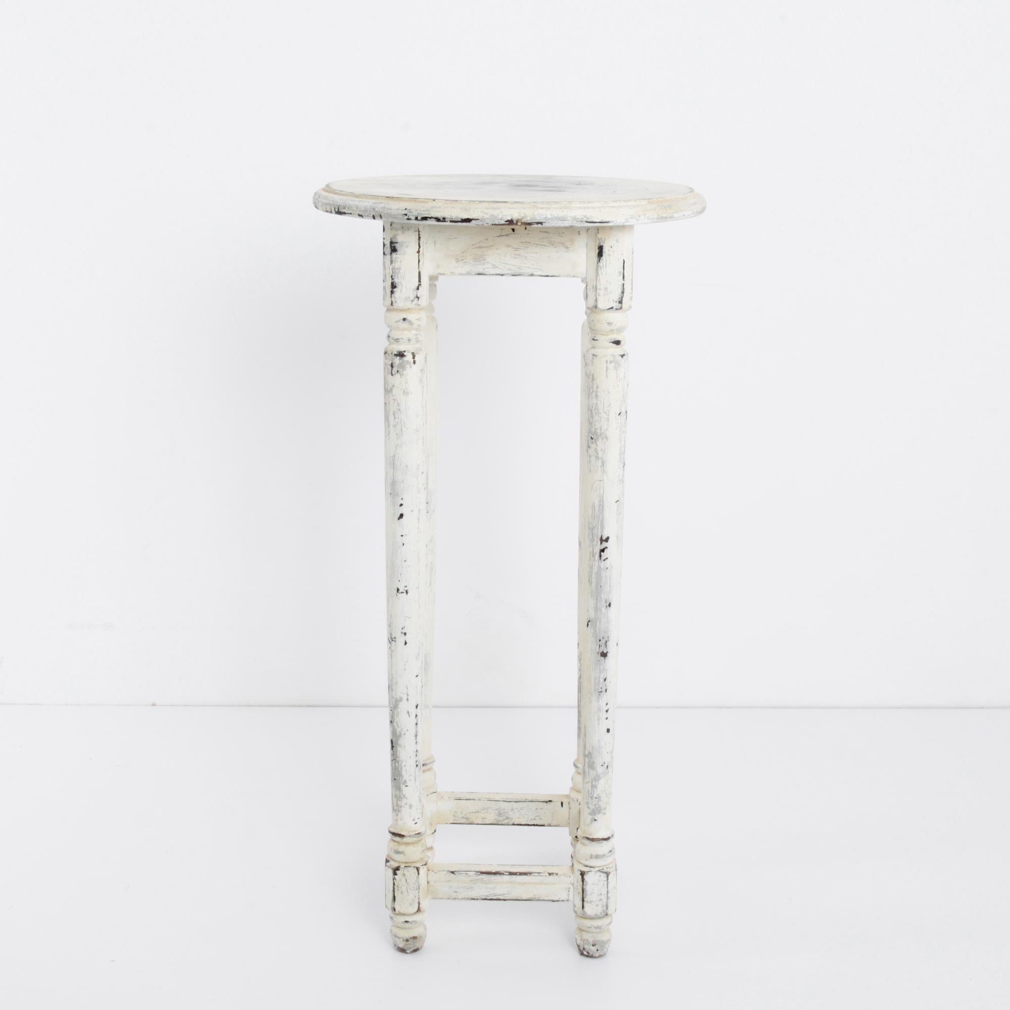 A white wooden side table from France, circa 1930. A small round tabletop sits atop a square apron; four turned legs descend to a square frame at the base. This interplay of shapes lends a pleasing variation to the tall, slender silhouette. The