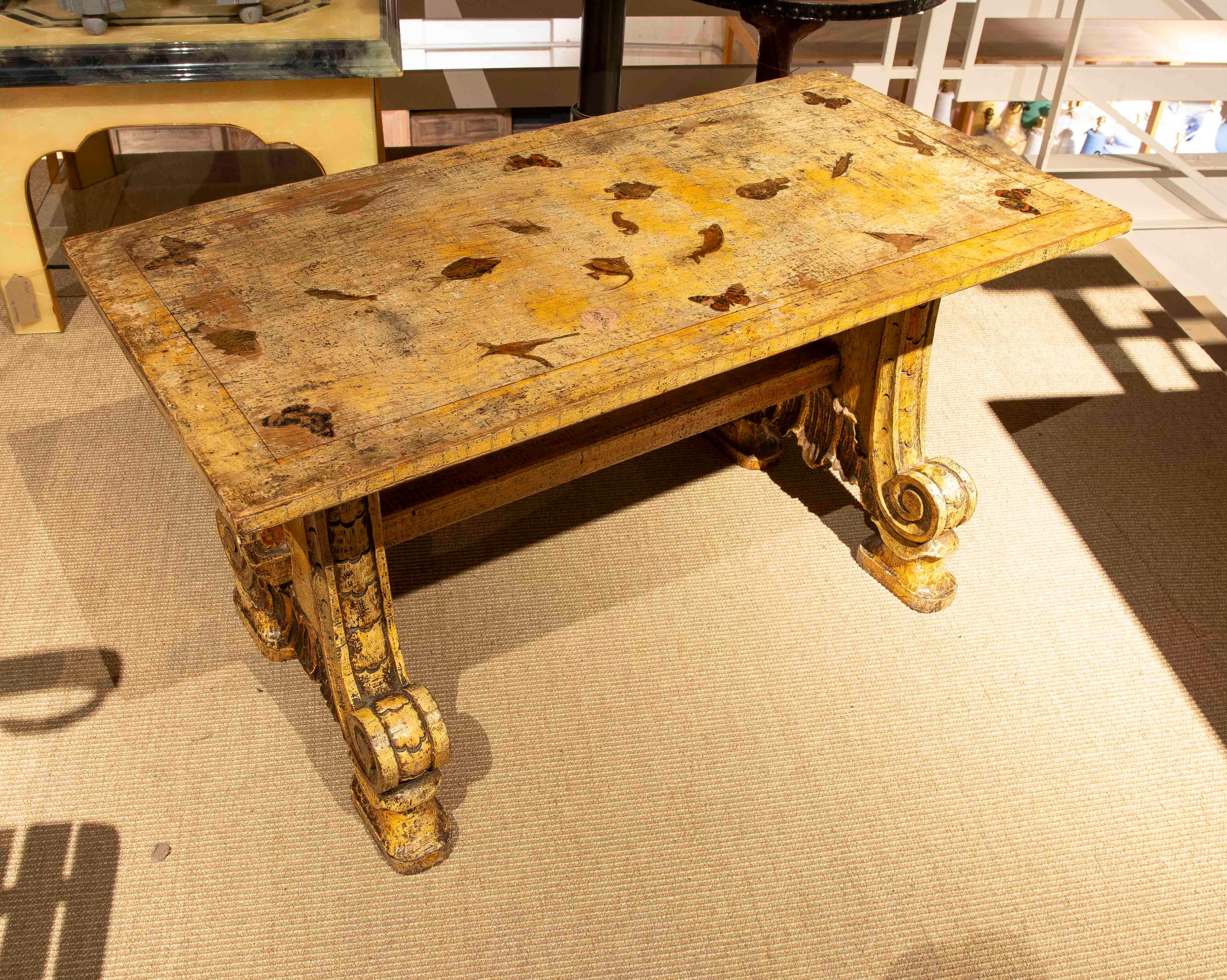 Majestic table en bois massif avec une finition vieillie qui met en valeur son caractère historique et sa beauté rustique. Son design imposant se caractérise par un plateau rectangulaire aux détails peints et une base aux pieds sculptés en forme de