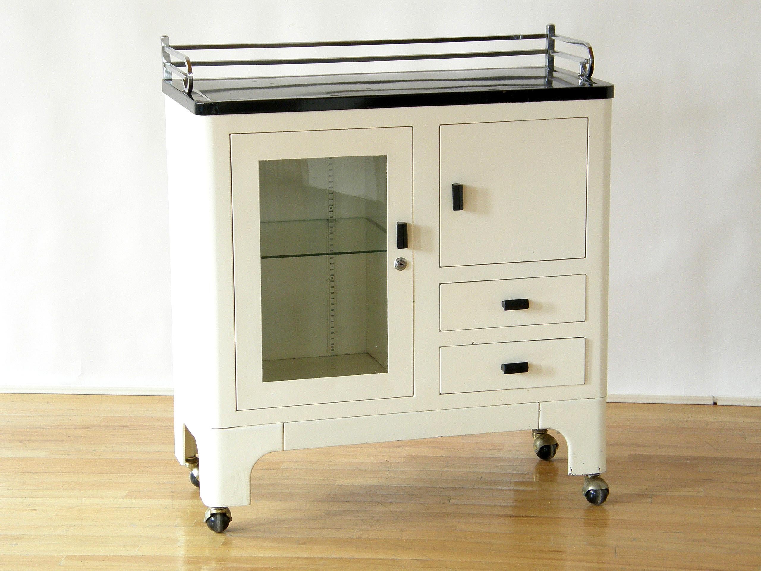 This circa 1930s medical cabinet has a machine age, modernist style with sleek rounded corners, a white enameled steel body, and a black enameled top with a streamlined, chrome-plated steel railing along the sides and back. The storage includes an