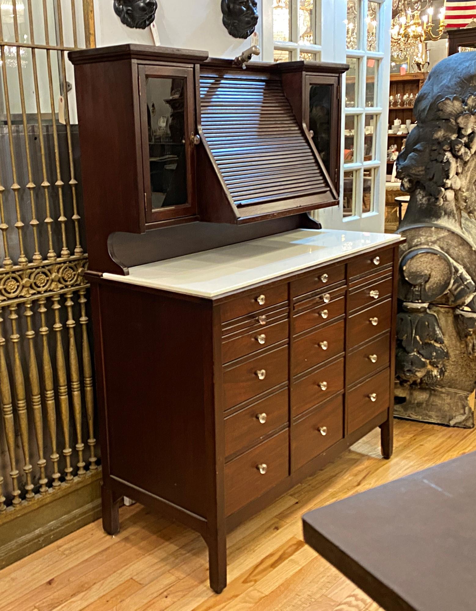 1930s dark tone mahogany ophthalmic or medical cabinet. Roll up center cover once held lenses for the eye doctor. Each side flanked by glass doors with two shelves each. This has lots of drawers and cubbyholes with a white glass counter top.