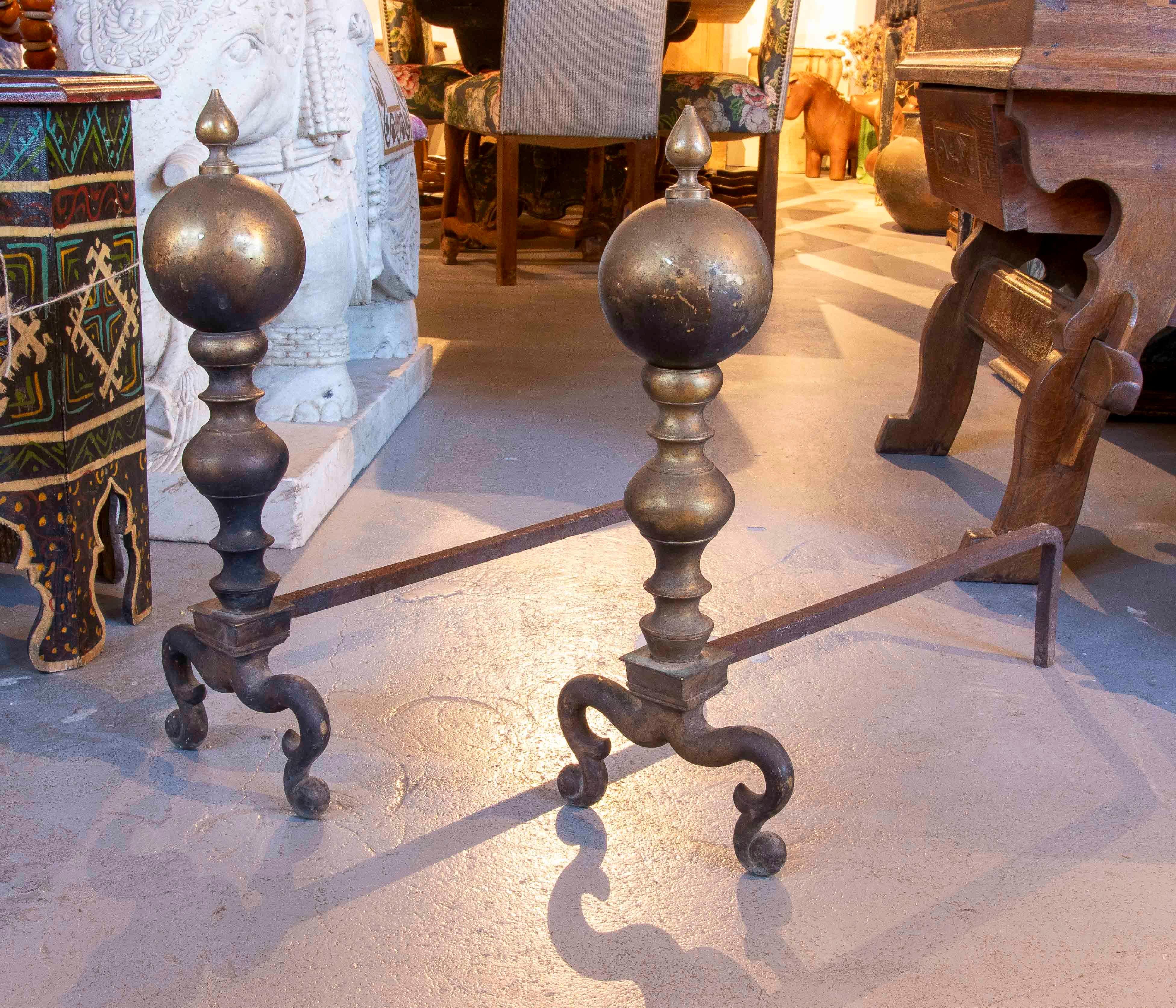 An elegant pair of fireplace andirons crafted from turned bronze, featuring pointed finials and a classic silhouette that combines intricate curves with a sturdy tripod base. They retain a beautiful original patina that enhances their historical