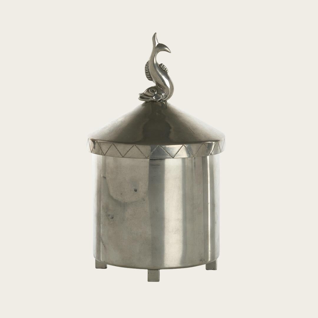 1930, Sweden. Swedish Grace lidded jar with detailed engraving of a ship with sail. Jar lid is crowned with a decorative dolphin. With feet and decorative engraving designs throughout. Marked Ystad Tenn and with Athena marking on underside.