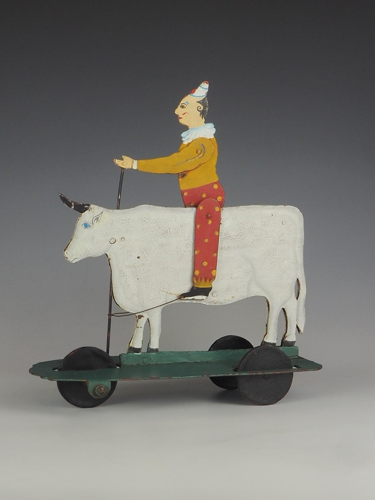 Circa 1930 double sided metal pull toy of a rodeo clown sitting on a bull with articulated arms and legs that move when the wheels turn, all in multi coloured original paint