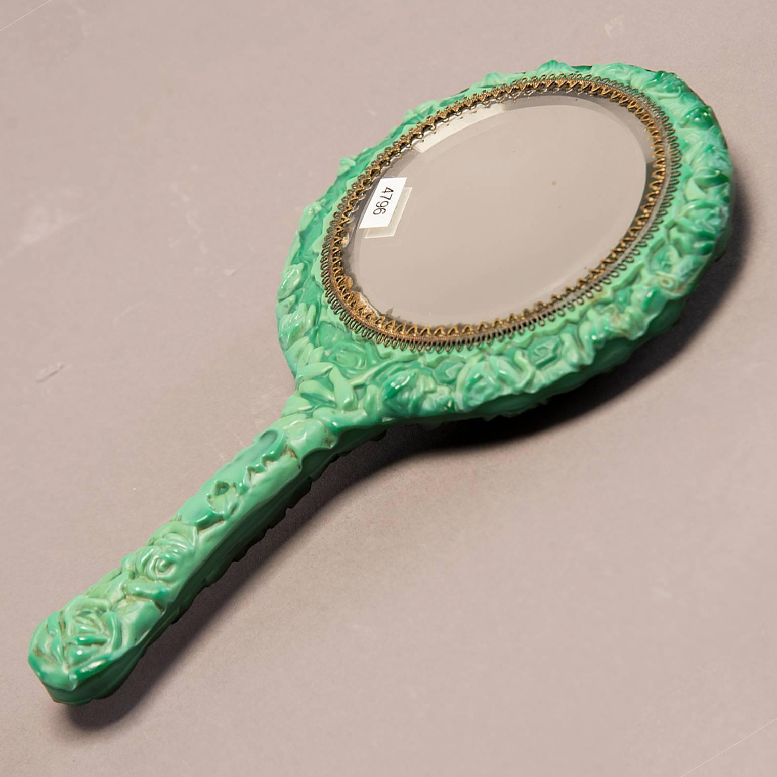 Deco era malachite glass hand mirror by Schlevogt from the Ingrid Collection. Malachite glass surround features a seated nude woman with flowing hair rendered in relief encircled by roses. Round, beveled edge mirror with brass filigree edge. Made in