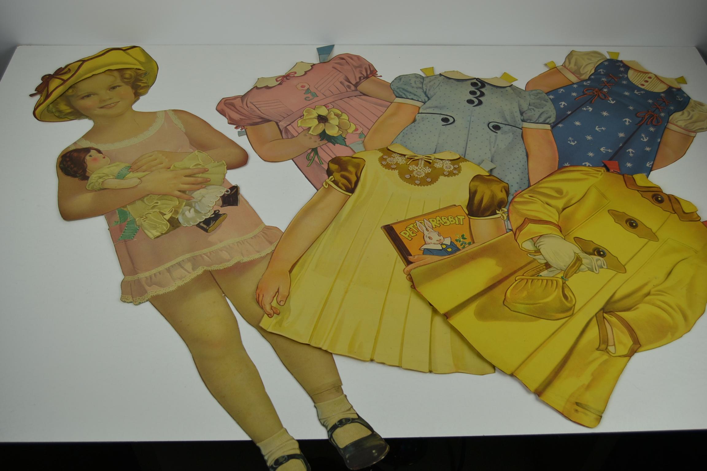 1930er Shirley Temple Papierpuppen-Set - große Größe 34 Zoll - 86 cm.
Shirley Temple trägt ein rosa Kleid, hält eine Puppe im Arm und trägt schwarze Schuhe.
Sie besteht aus 2 Teilen: dem Oberkörper und den Beinen.
Du kannst ihre Kleidung wechseln,