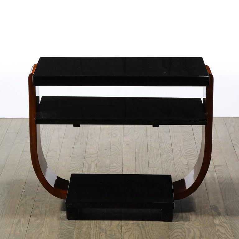These elegant Art Deco Machine Age side tables was realized in the United States circa 1935. They feature curvilinear walnut legs connected to a black lacquer base which supports the lustrous black lacquer two tier top. With their clean modernist