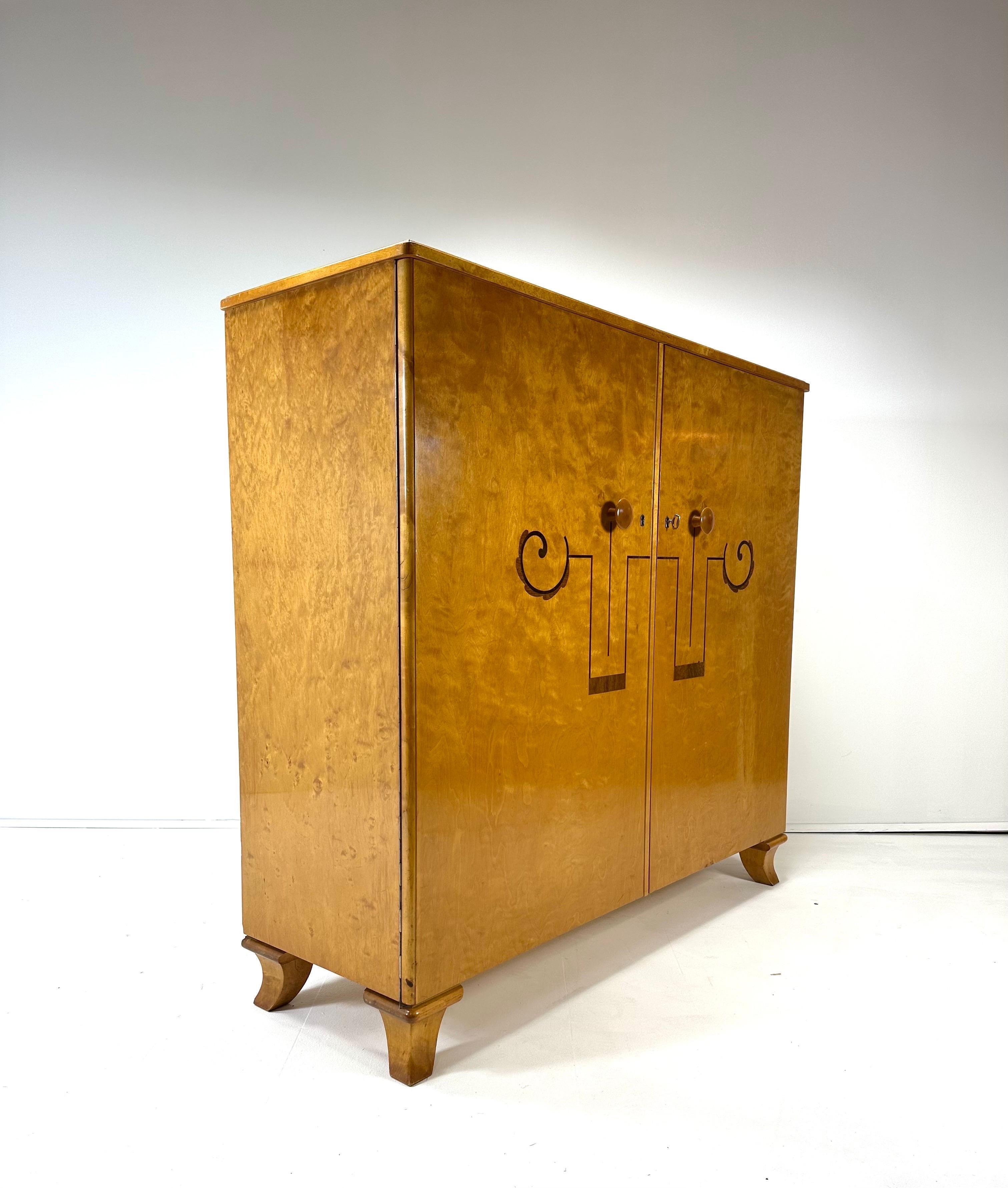 Armoire suédoise art déco des années 1930. Meuble en bois de bouleau avec incrustations décoratives. Deux portes dissimulent des étagères intérieures réglables et deux petits tiroirs. Clé incluse