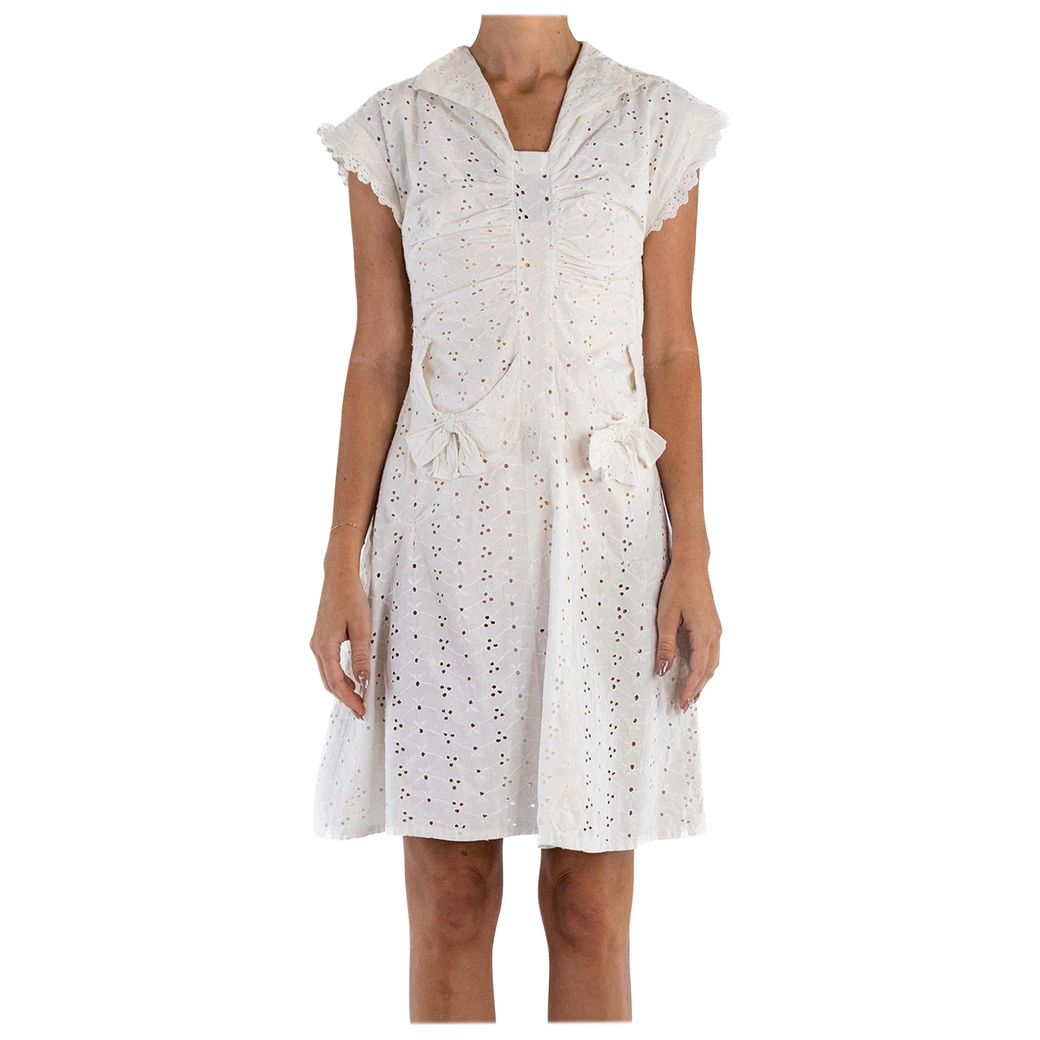Petite robe en coton blanc à œillets et dentelle avec poches à nœud, années 1930